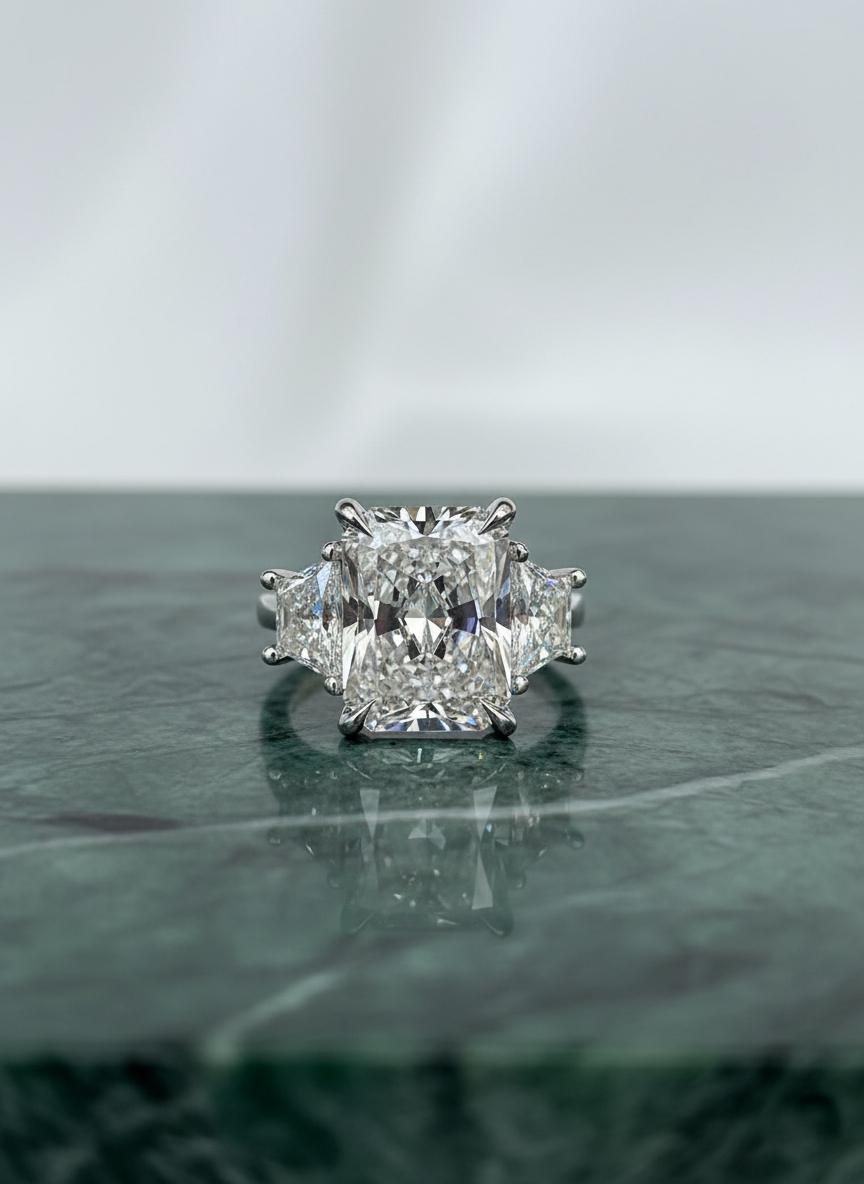 Un spectaculaire diamant radiant de 4 carats certifié GIA, de couleur E et de pureté VS2, occupe le devant de la scène dans cette bague intemporelle en platine à trois pierres. Le diamant affiche une brillance et un feu remarquables, parfaitement