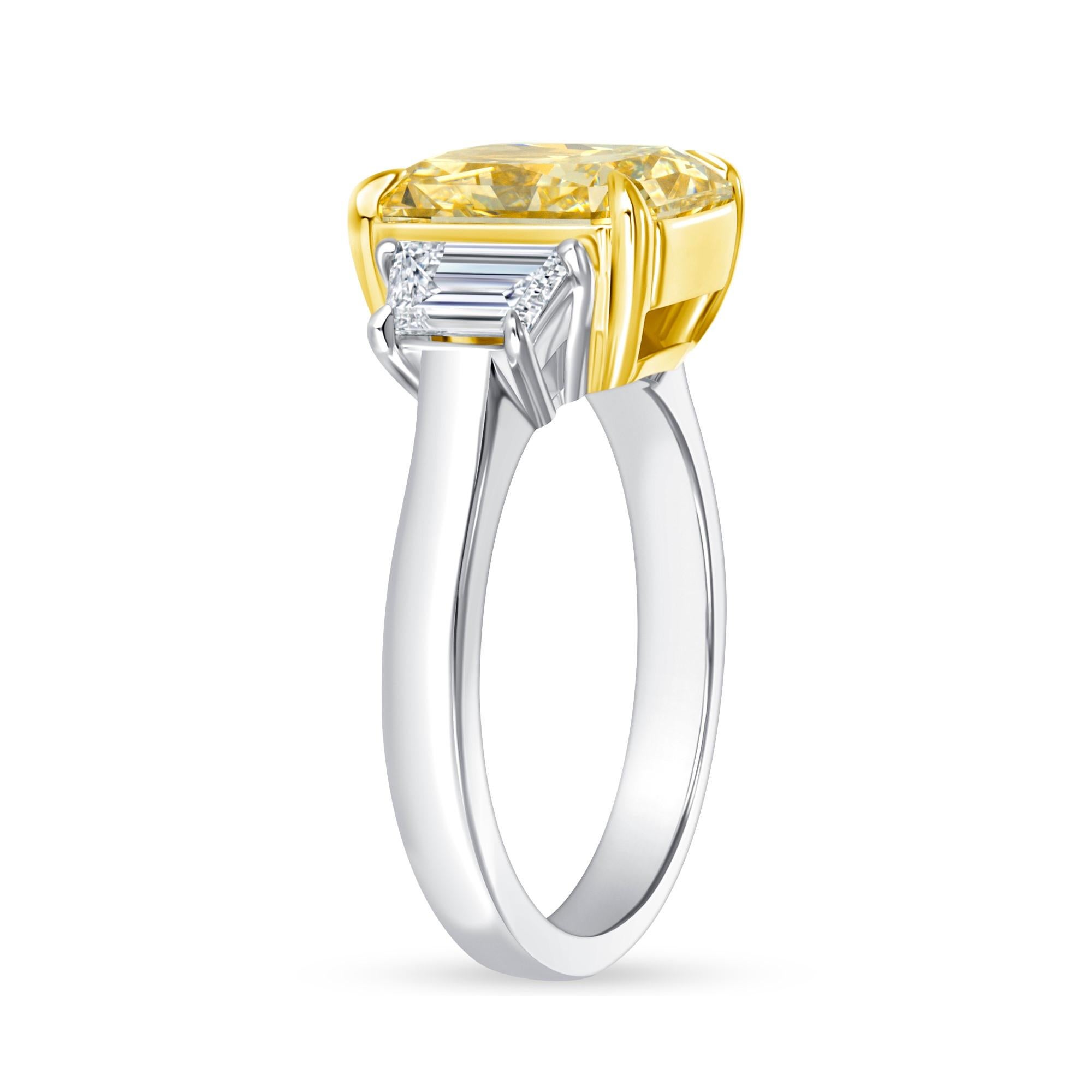 Este anillo de tres piedras tiene un diamante de talla Fancy Yellow Radiant de 4,12 quilates con certificado GIA de claridad VS1 y  dos diamantes de talla trapezoidal escalonada certificados por el GIA, de color D y claridad VVS2, engastados en oro