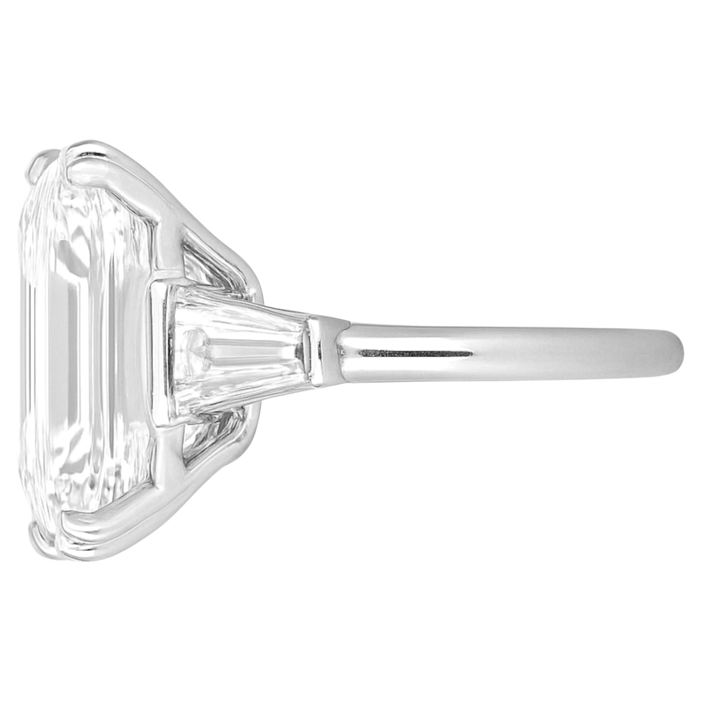 Questo magnifico anello di fidanzamento con tre pietre ha una presenza imponente grazie al suo diamante taglio smeraldo da 5 carati, montato in un'elegante montatura in oro bianco 18 carati. Accentuato da due diamanti baguette affusolati, il design