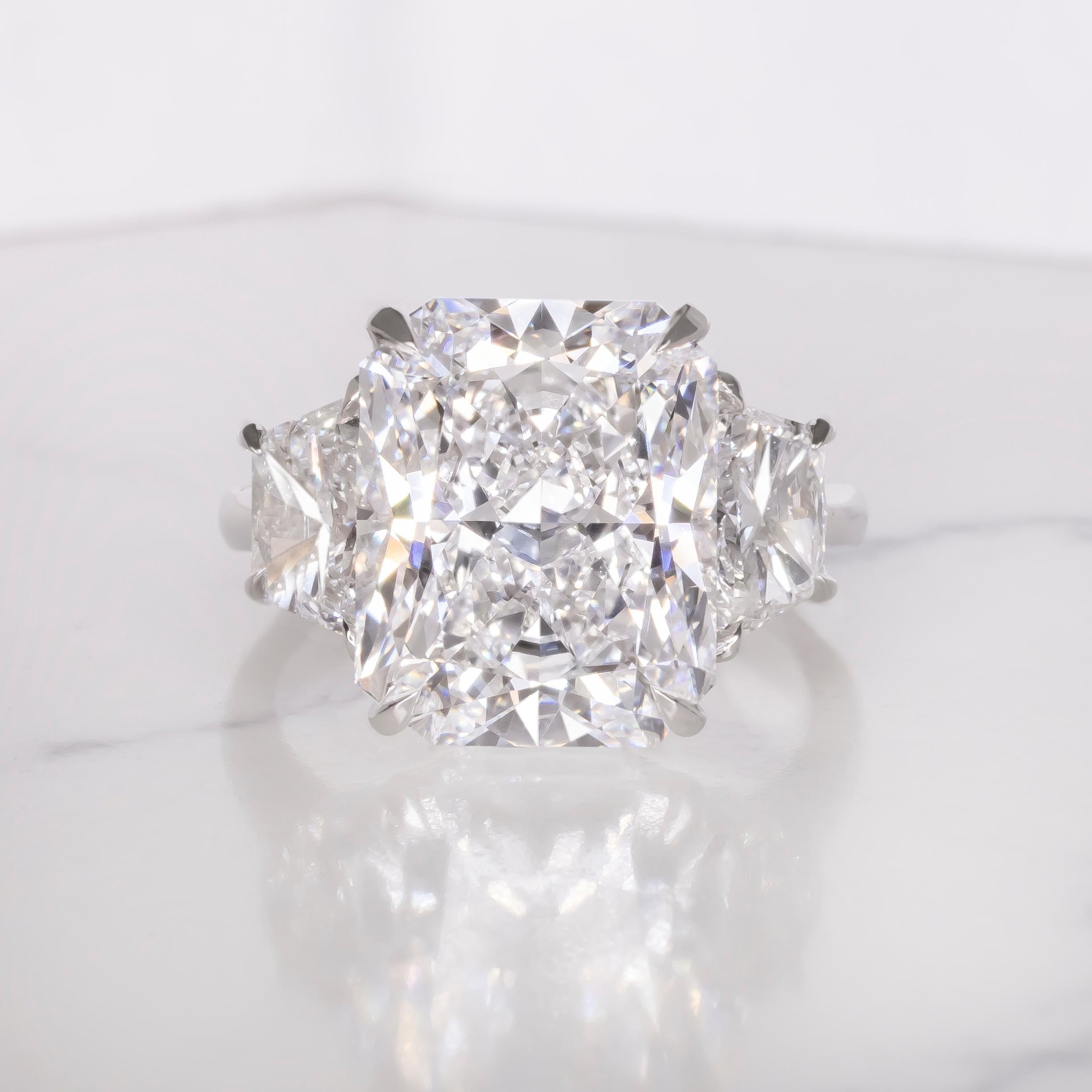 This striking engagement ring showcases a GIA certified 5 carat radiant cut diamond set in platinum, offering a bold yet refined silhouette. Flanked by two trapezoid diamonds, the three stone design adds dimension and visual harmony while drawing