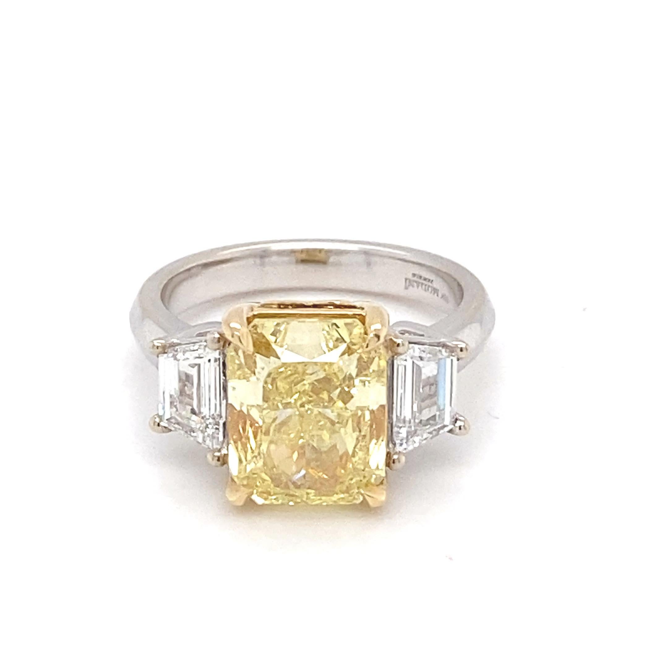 This magnificent ring boasts a Gia Certified 5.01 Carat Intense Fancy Yellow cushion diamond as the center stone with two trapezoid white diamond as side stone. The center stone is mounted in yellow gold whereas the side stone and the shank is in