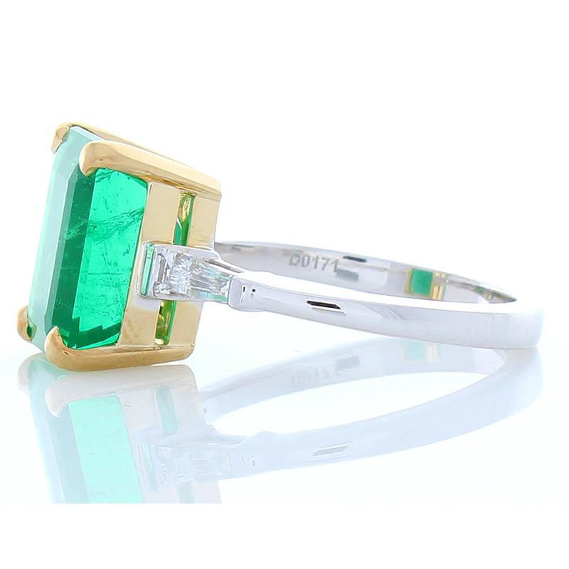 This is a three-stone ring that is timeless, classic, and richly unique. A 5.26 carat octagonal cut green emerald is carefully prong set in the center in shiny yellow gold accenting and measures 11.61x8.46x6.99MM. The gem source is Zambia; its color