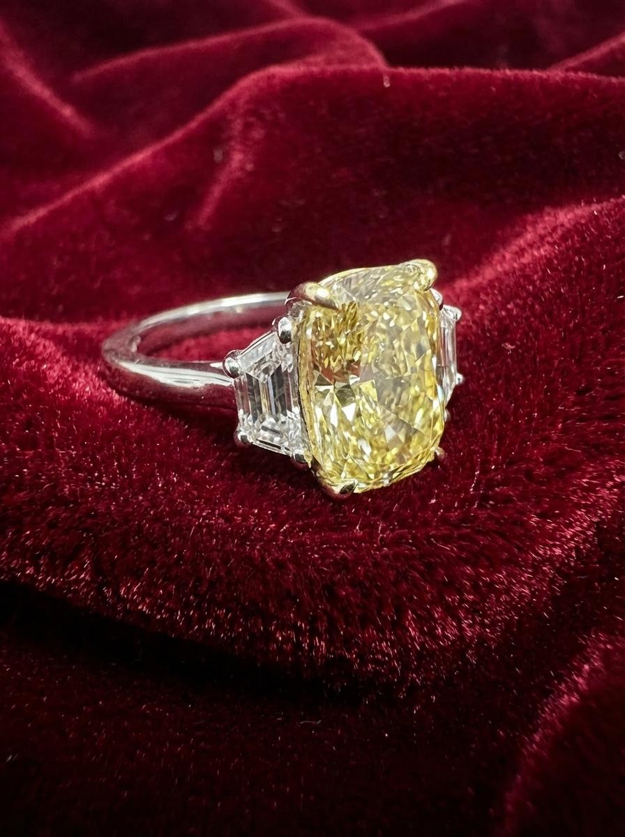 Este exquisito anillo de tres piedras tiene un total de 5,80 quilates de diamantes excepcionales, todos certificados por el GIA. En su centro descansa un llamativo diamante brillante modificado en cojín Amarillo Fancy de 5,00 quilates, que muestra