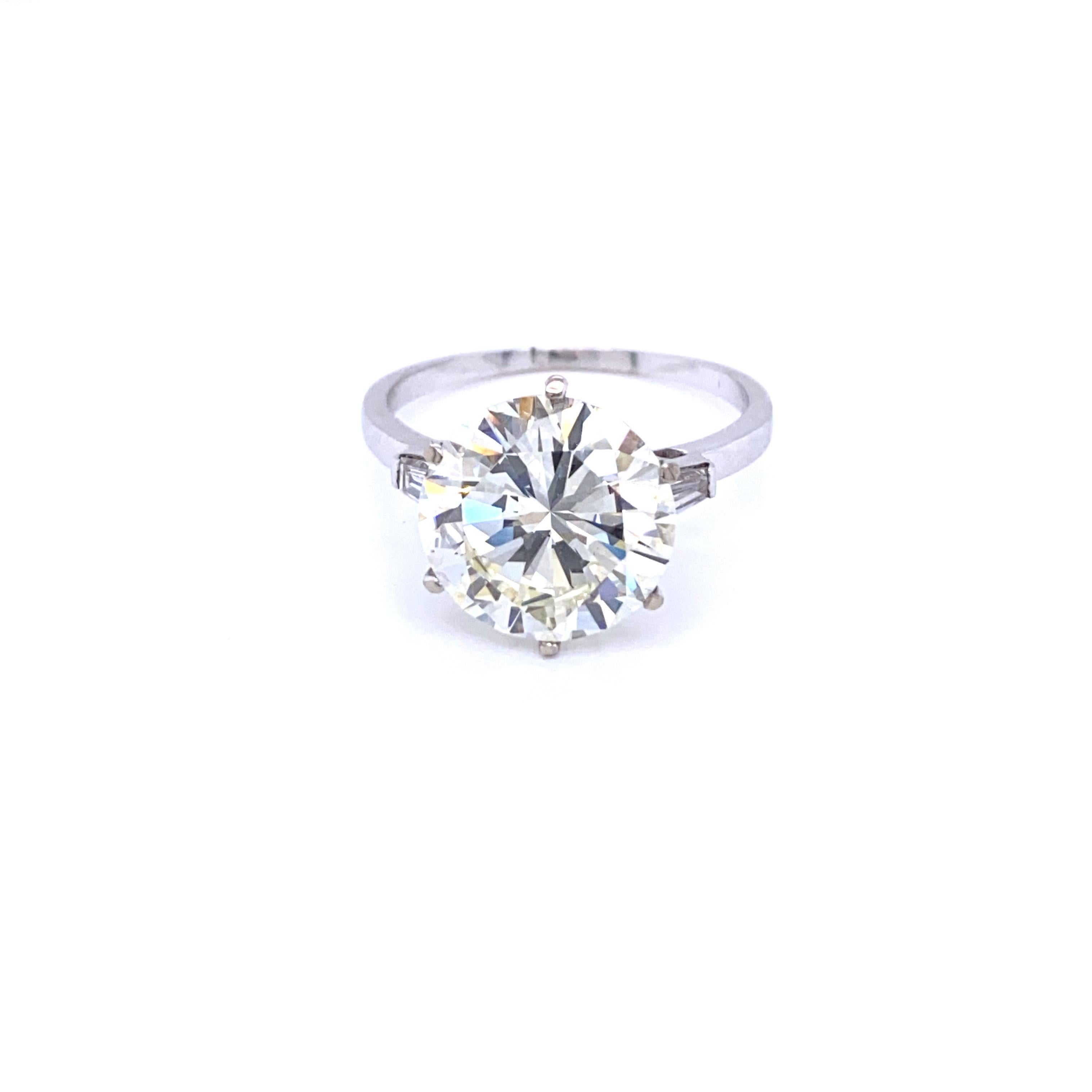 An exceptional estate engagement ring set in 18k white Gold, featuring a large 5.87 ct Round brilliant-cut Diamond, graded M Color Vvs2 clarity, Very Good proportions and faint fluorescence, surrounded by 0,30 carats of baguette cut diamonds.

The
