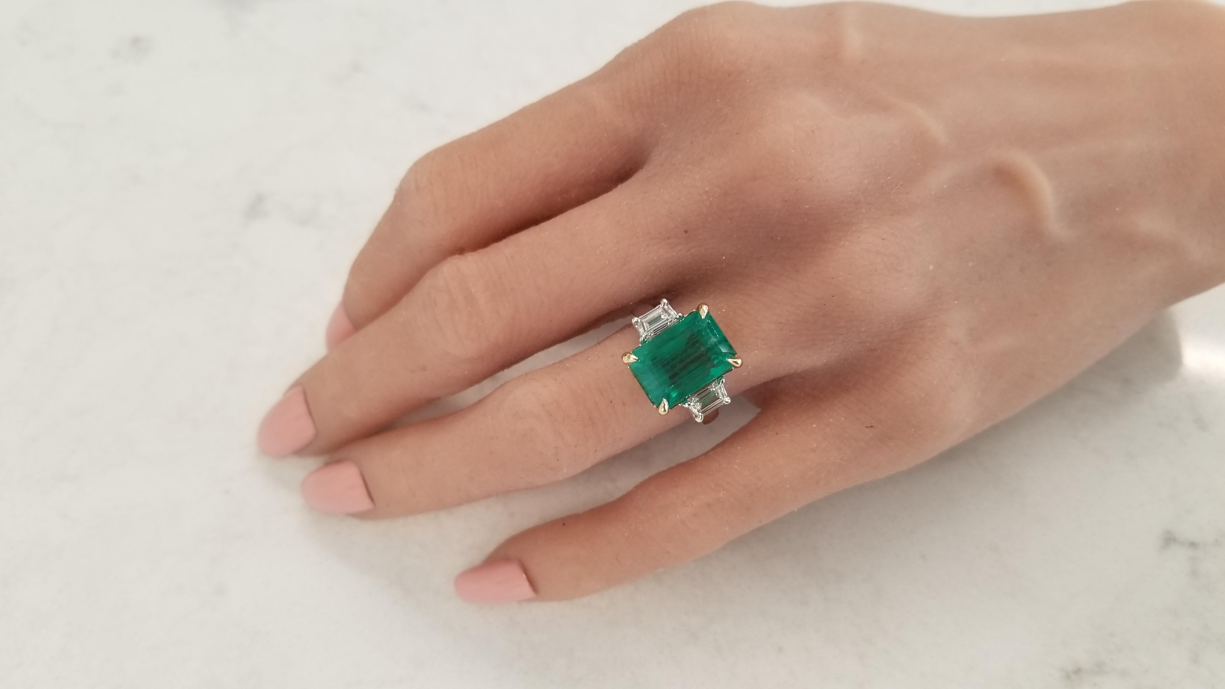 This is a three-stone ring that is timeless, classic, and richly unique. A 7.80 carat octagonal cut green emerald is skillfully prong set in the center in shiny yellow gold accenting and measures 12.50x9.00MM. The gem source is Zambia; its color is