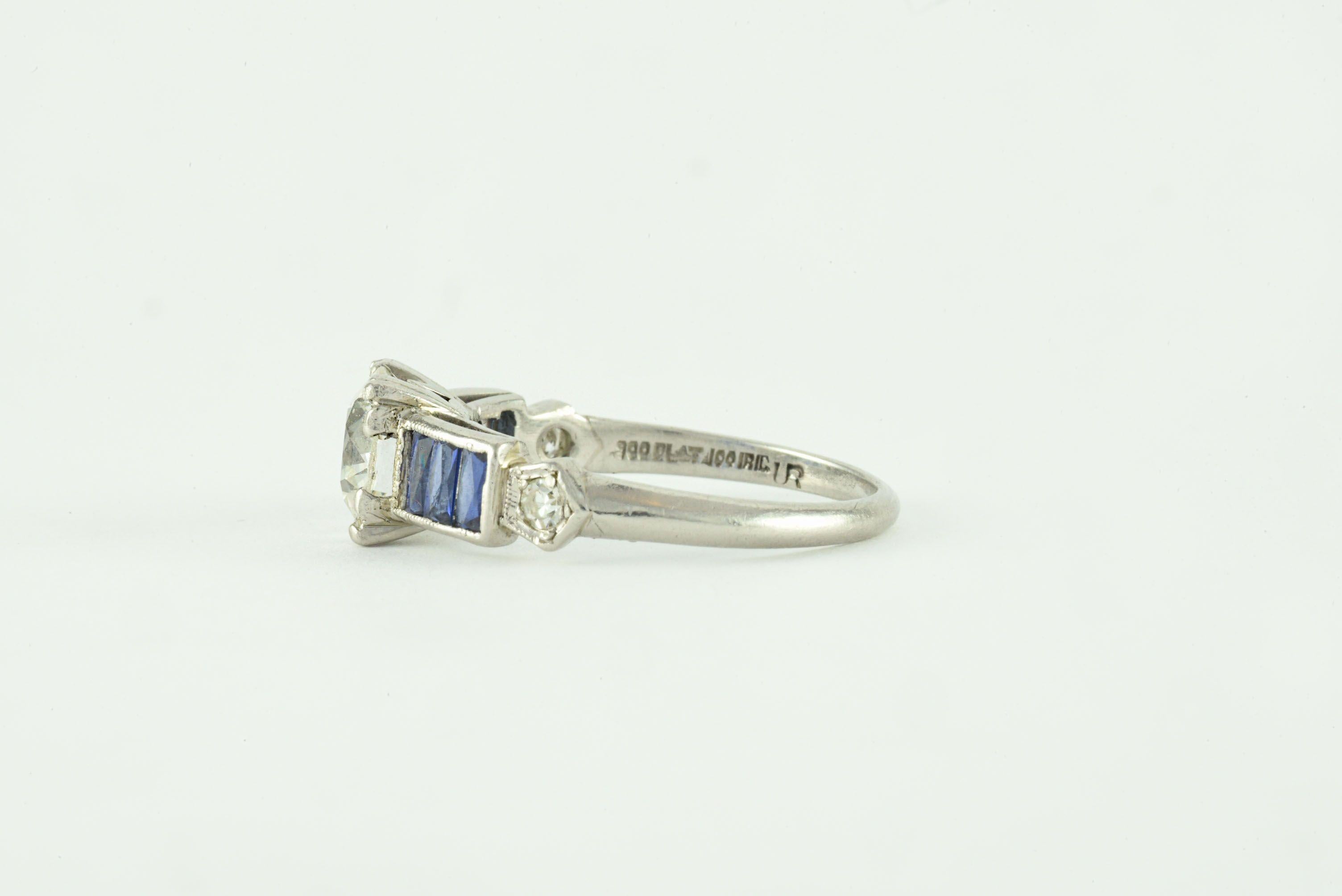 In this Art Deco gem, a circular brilliant cut diamond measuring 1.08 carats, J color, VS1 clarity sparkles in the center, flanked by six natural blue calibre baguette-shaped sapphires and two single cut diamonds totaling 0.07 carats, beautifully
