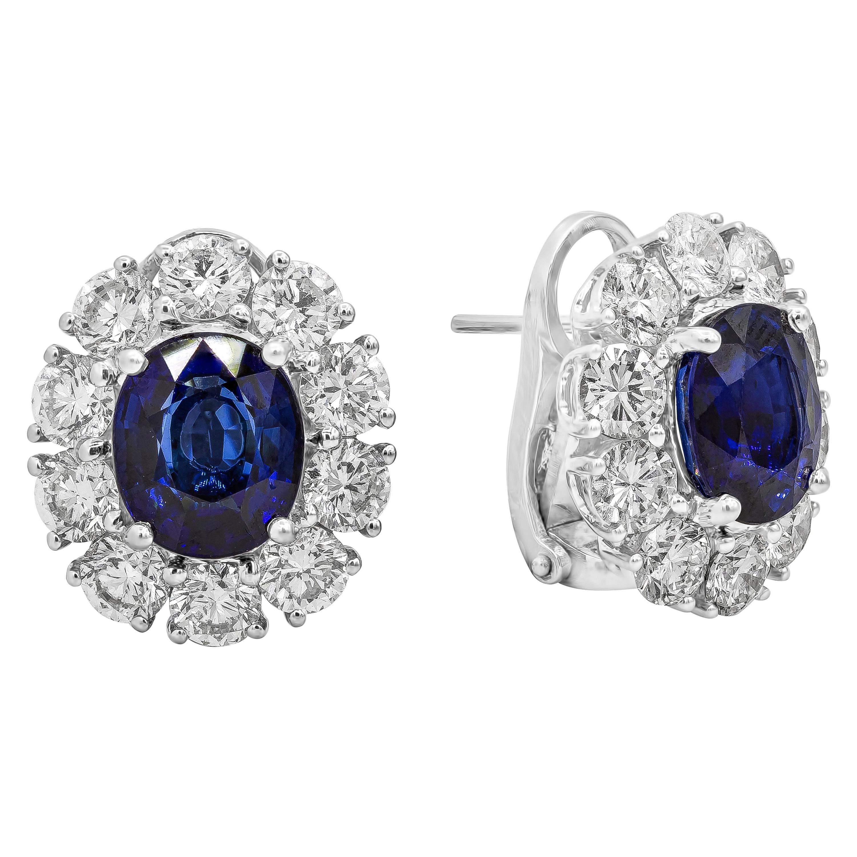 A unique pair of halo earrings showcasing two GIA certified heated blue sapphires from Madagascar weighing 6.05 carats total. Accented with a row of brilliant round diamonds weighing 4.27 carats total. Made in 18k White Gold. Omega Clip with