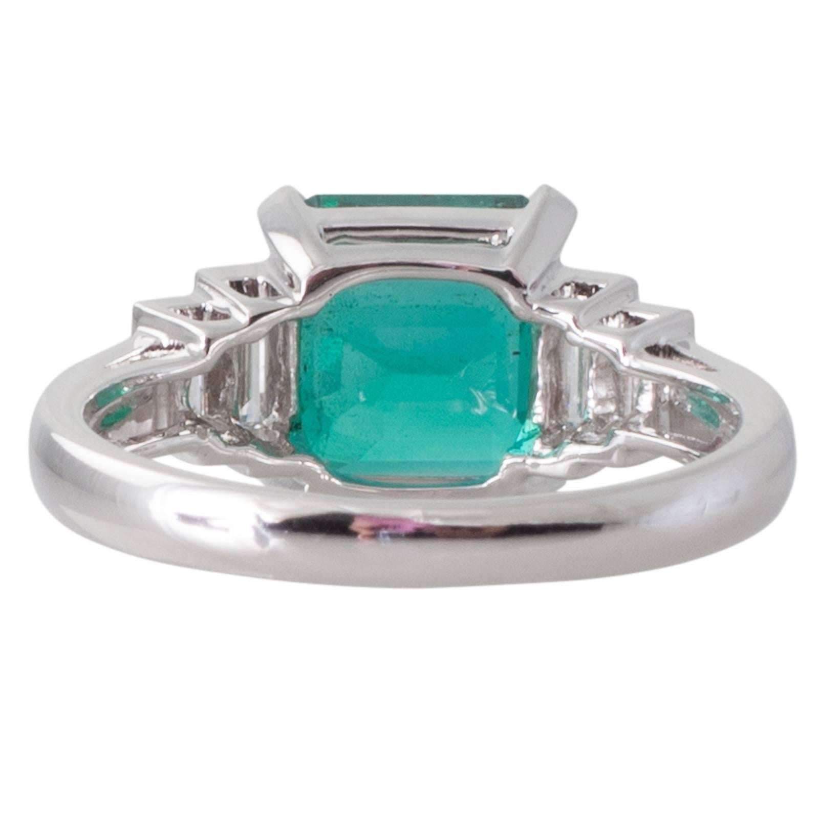 An 18ct white gold ring four claw set to the centre with a GIA certified 3.34ct Zambian octagonal step cut emerald above a railed gallery and to either side a pair of baguette cut diamonds to upswept shoulders and a plain polished band. Total