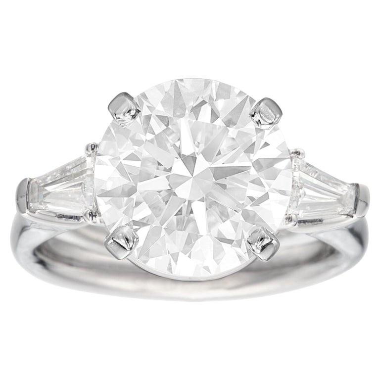 Adórnate con el epítome de la elegancia y la sofisticación con este Anillo de Diamantes de Corte Brillante Redondo Certificado GIA de 5 Quilates, acentuado por diamantes baguette cónicos. En el corazón de este exquisito anillo brilla un