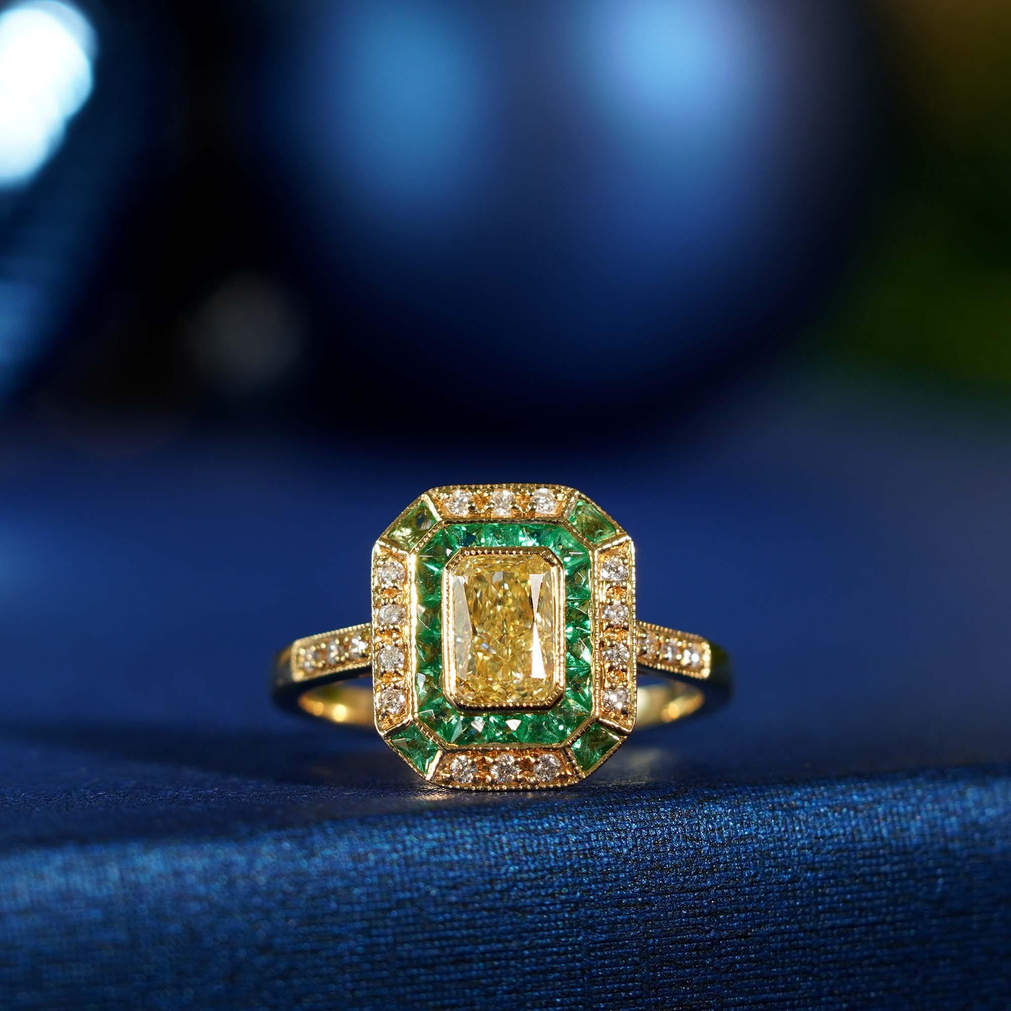 A fabulous Art Deco inspired ring stars a resplendent rich shiny fancy yellow emerald cut diamond weighing .82 carat. The glorious gemstone radiates from within an octagonal yellow gold bezel frame in bright green French cut emeralds, outlined in