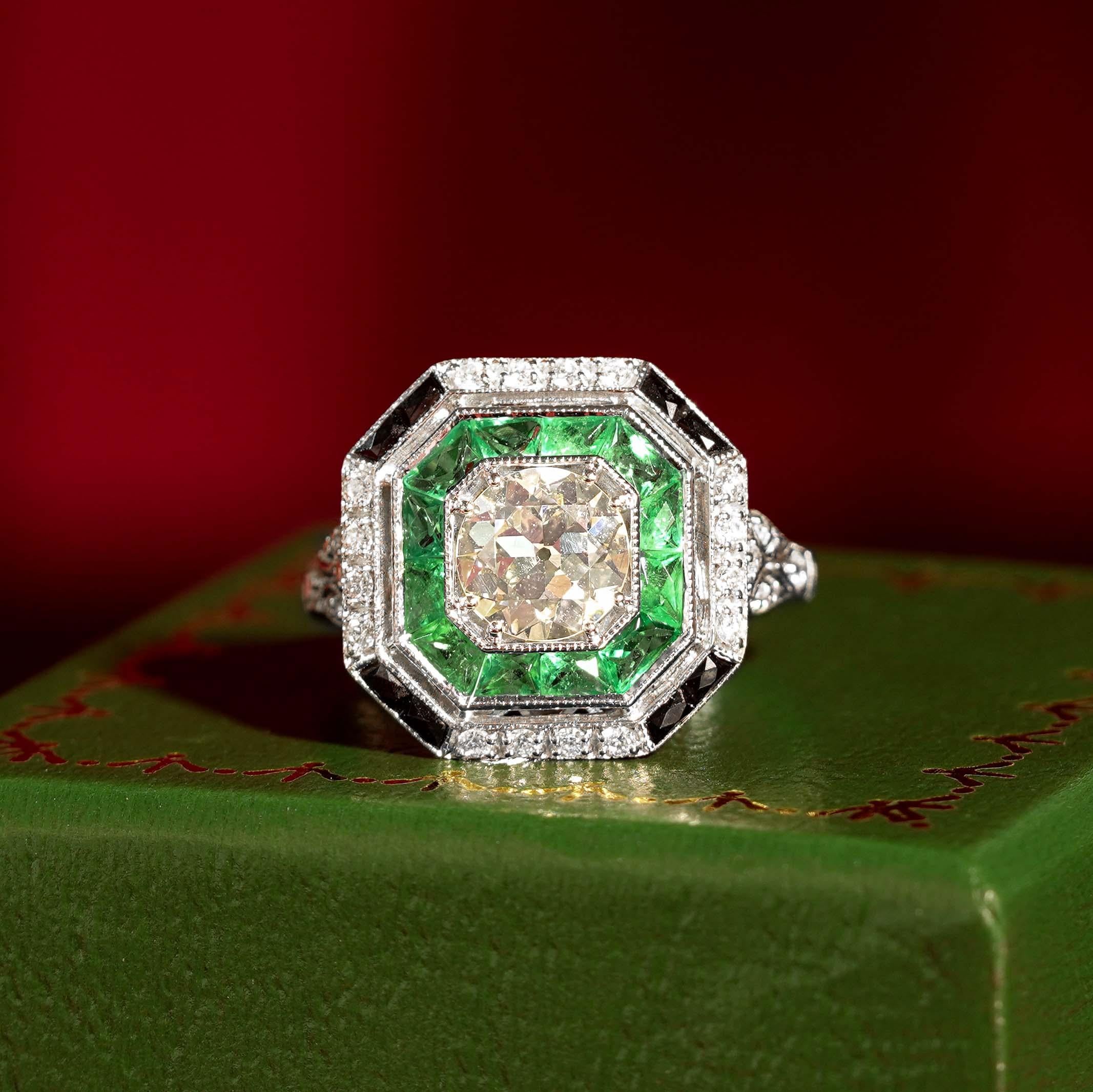 An 18k white gold Art-Deco design diamond emerald and onyx target ring, center diamond with an outer ring of round diamonds an inner one of French cut emeralds and onyx and finished with millgrain edging, finished with diamond shoulders.

Ring