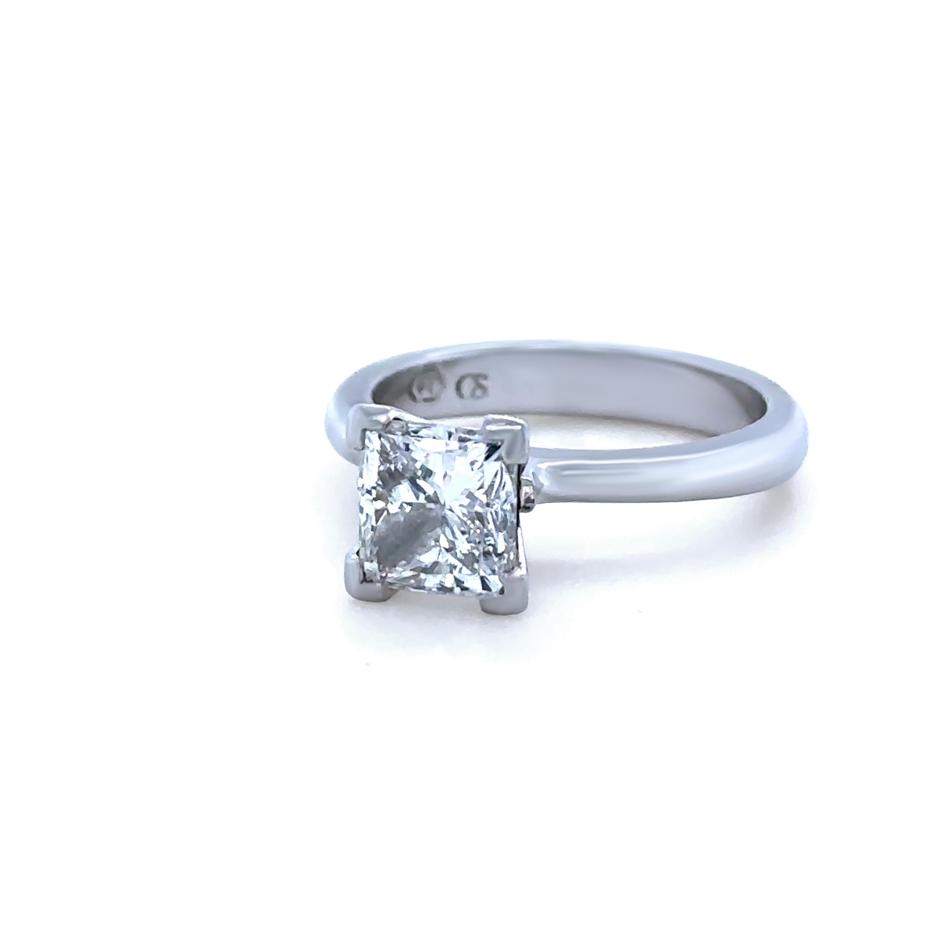 Un intemporal anillo de compromiso de platino con un impresionante diamante talla princesa de 1,56 ct, elegantemente engastado en un seguro engaste de cuatro garras en forma de L sobre una banda pulida suavemente ahusada. El diamante central tiene