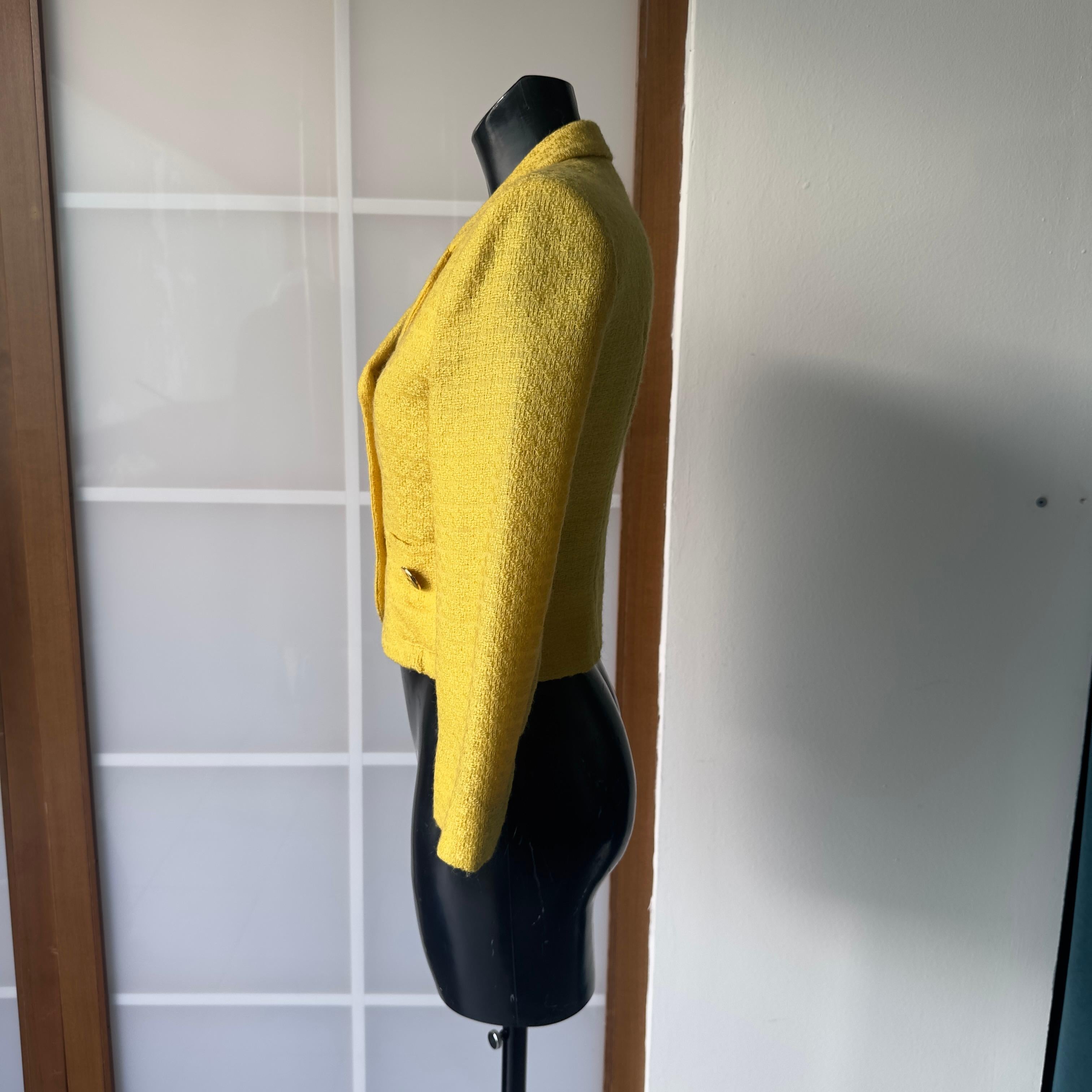 Un raro pezzo d'archivio firmato Moschino Cheap and Chic, risalente agli iconici anni '90: questa giacca corta in lana bouclé gialla è un perfetto esempio dello spirito ironico e sofisticato del brand. Il dettaglio distintivo? I bottoni dorati a
