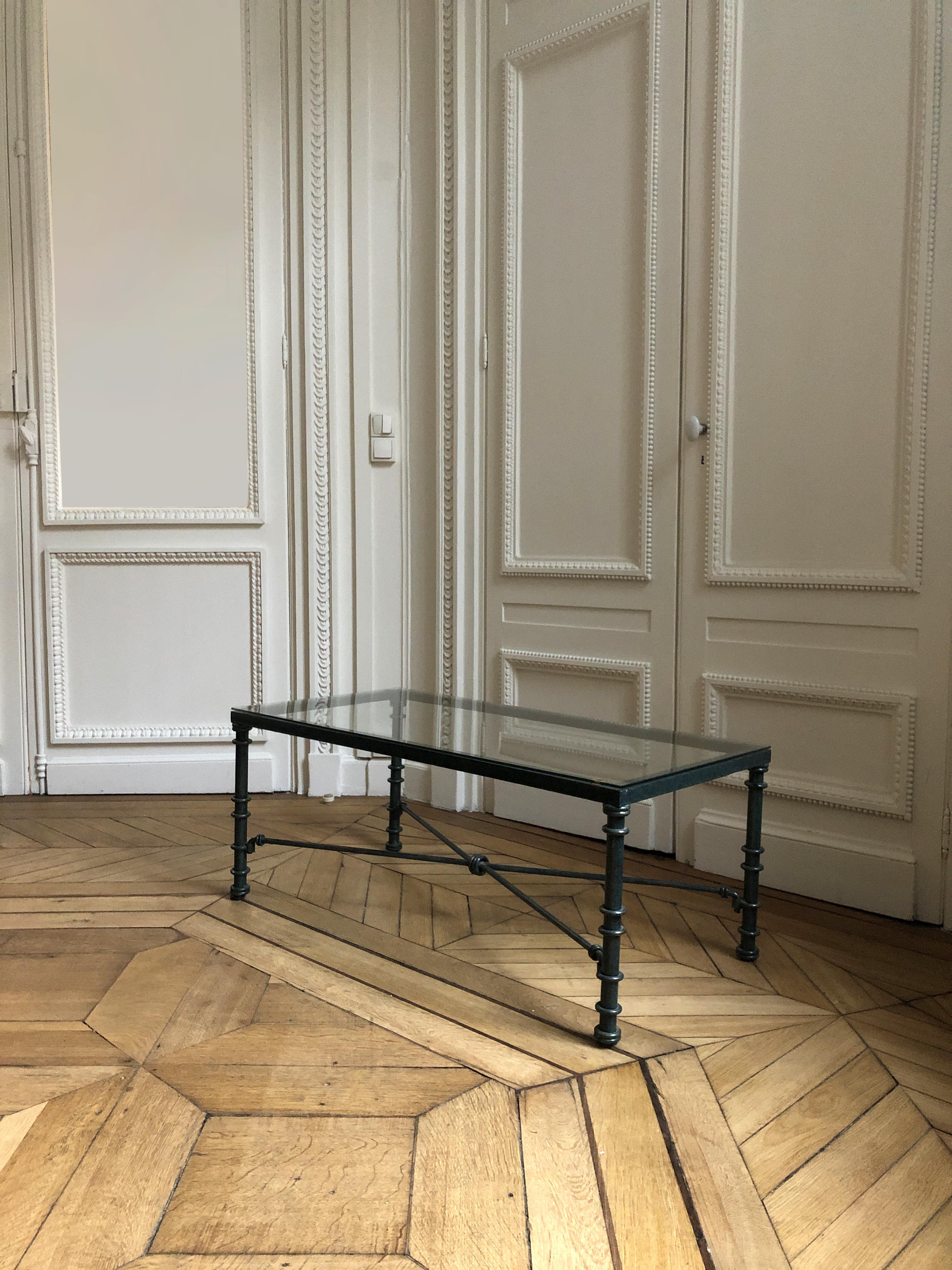 Nice coffee table with a glass top resting on a wrought iron base. The base has a dark green-grey patina. It has ringed uprights joined by an X-shaped spacer. The glass top is hold with suction cups. 
It is a bit hard to date this around 1950 in