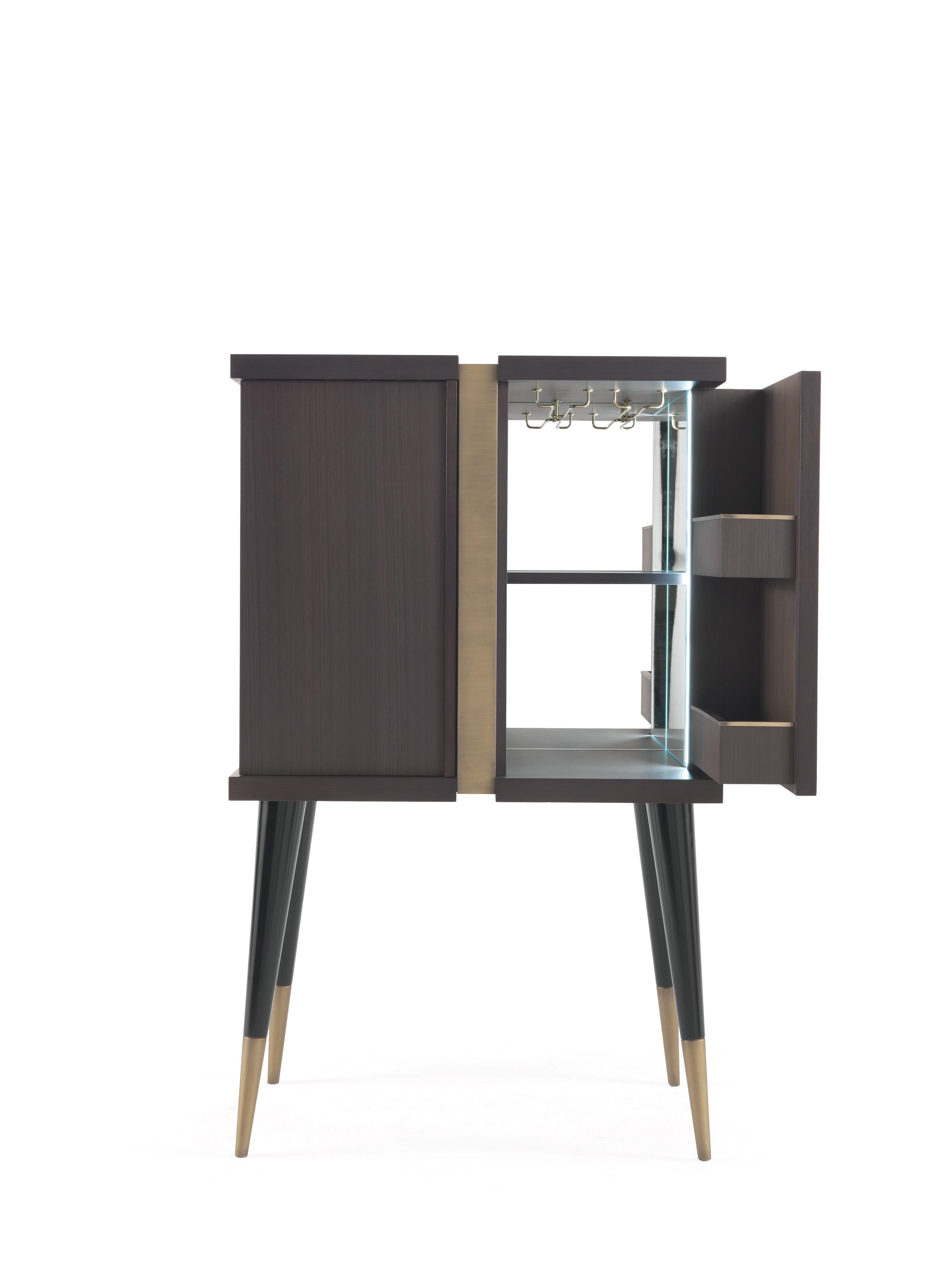 Featuring a solid structure in Tay wood tinted smoky grey with a bronzed central band, the Sean bar unit reveals a luminous interior with mirror and polished brass profiles, while the inclined legs with brass ferrule add a vintage touch to the piece