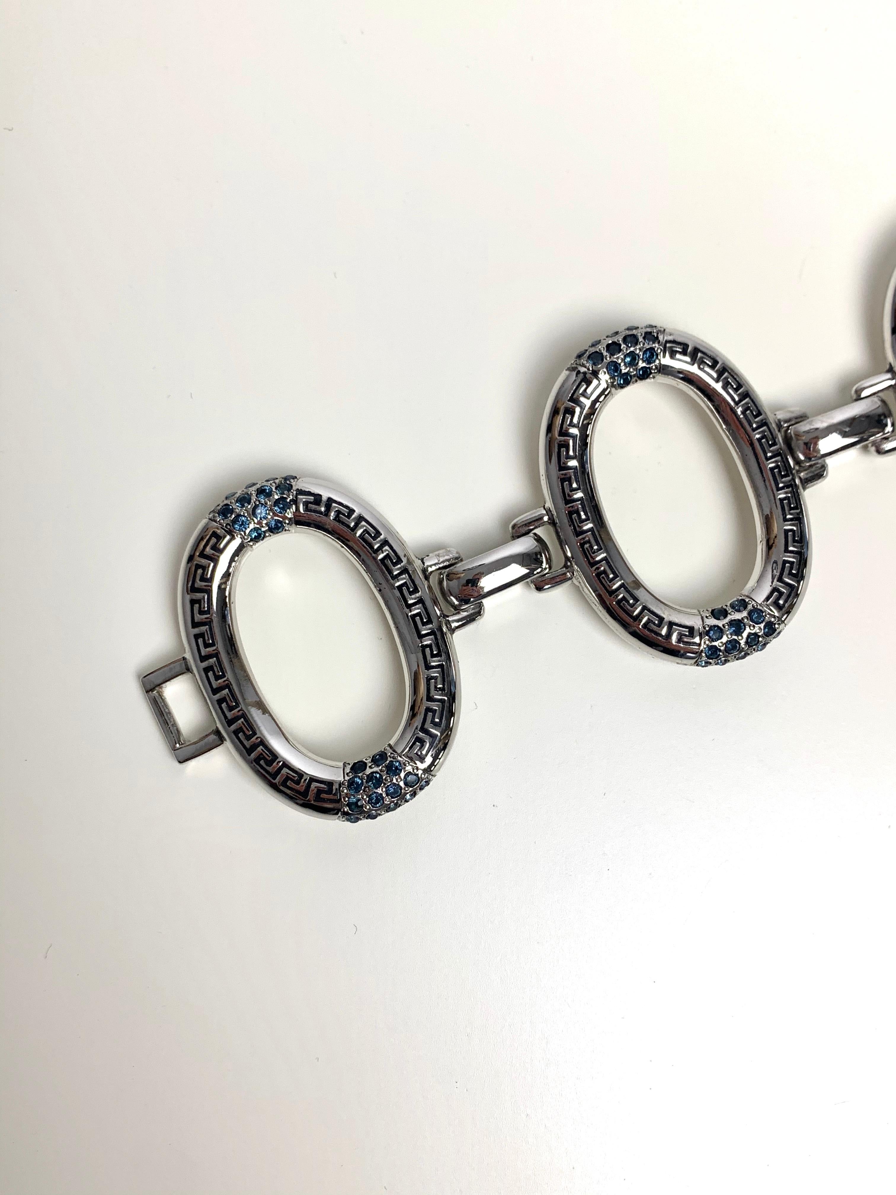 Dieses ovale Armband von Gianni Versace besteht aus großen, ovalen Gliedern, in die das Motiv einer griechischen Fliese eingraviert ist. Sie haben auch dunkelblaue Steine am oberen und unteren Ende, die einen schönen Kontrast zum silbernen Armband