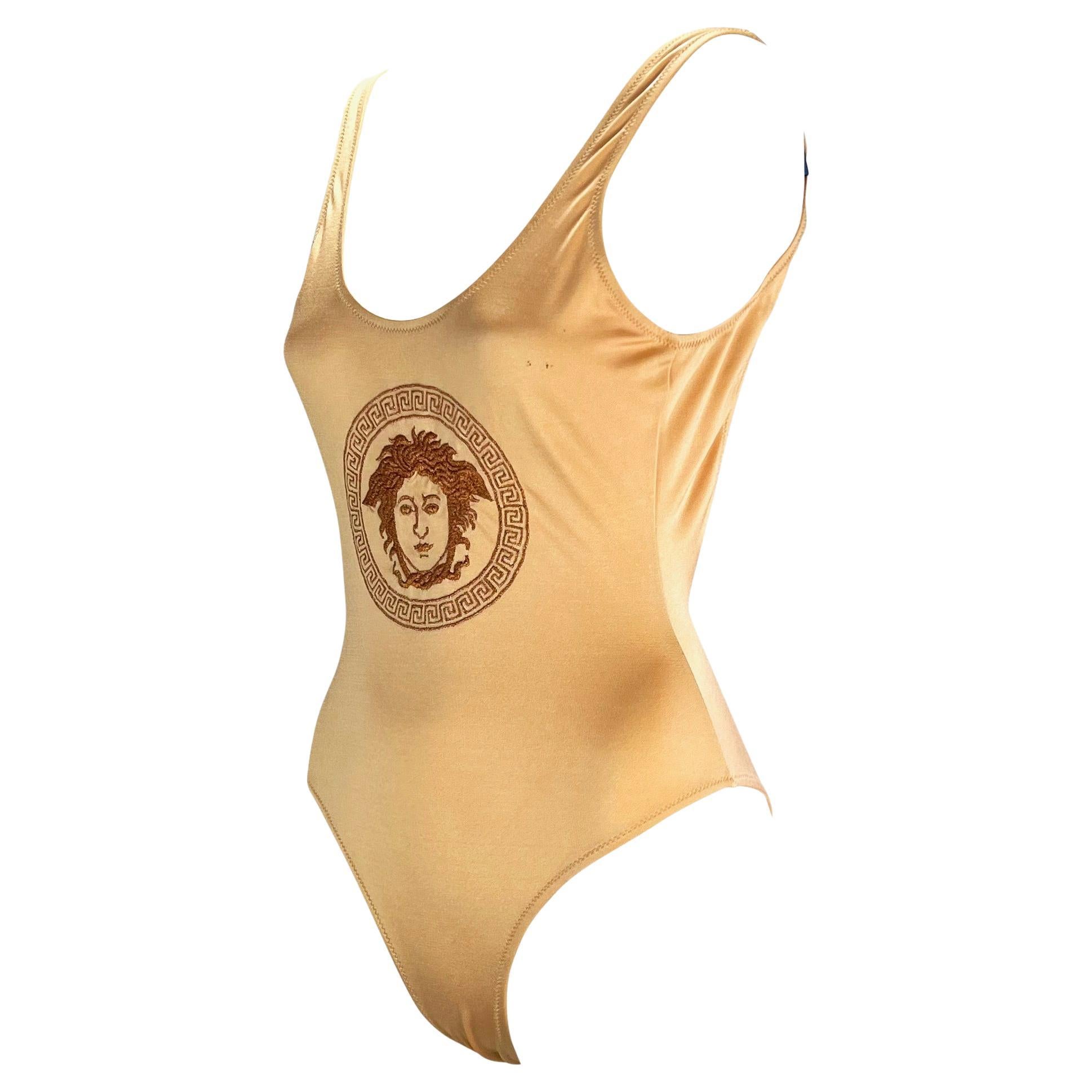 Presenting a beautiful beige Gianni Versace one piece bathing suit. From the 1990s, this shimmery beige swim suit proudly features an embroidered Versace Medusa with surrounding Greek key at the front center. The piece features a scoop neckline and