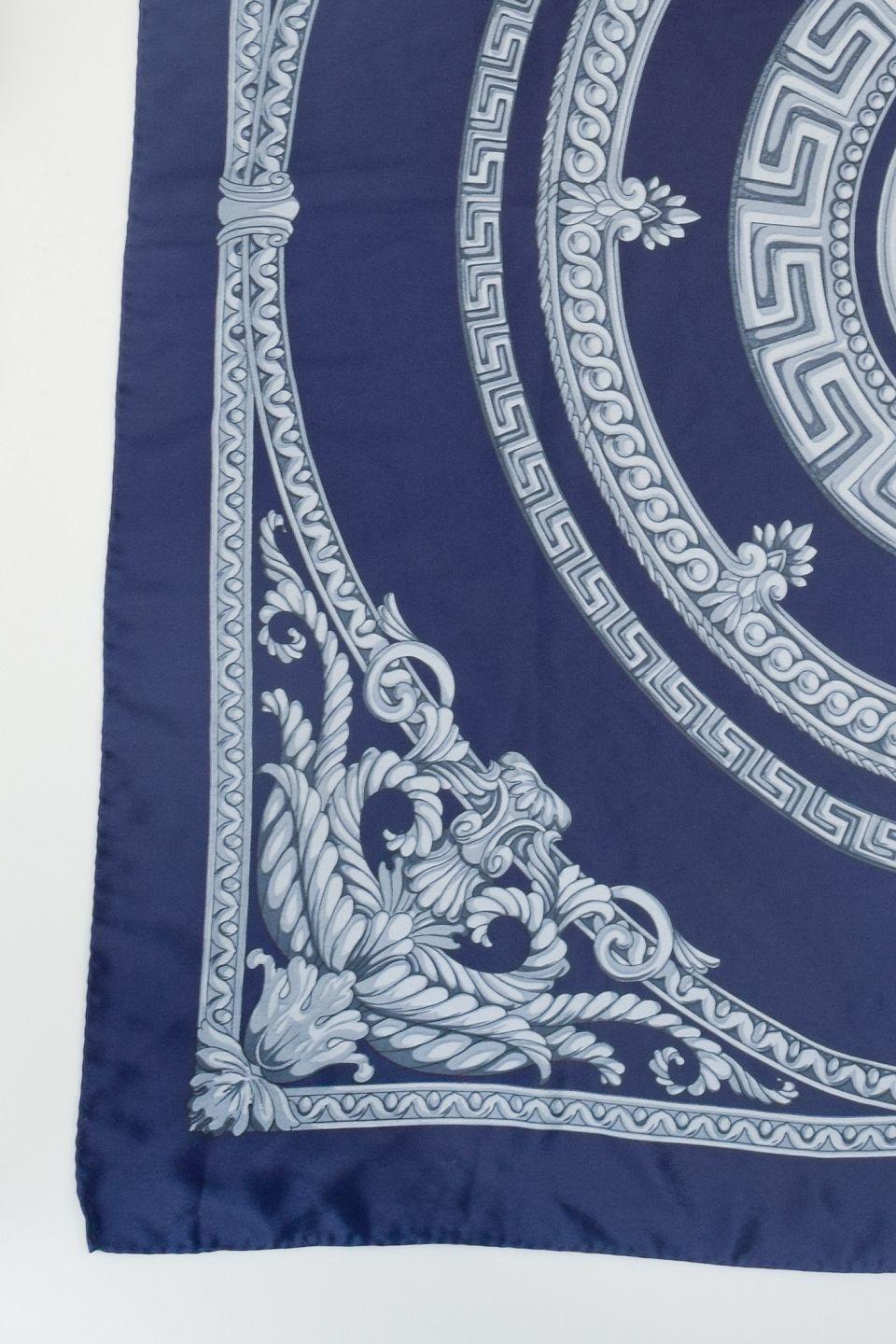 Esclusivo foulard in pura seta di Gianni Versace. 
Disegnato in blu profondo con l'iconico motivo della Medusa al centro, incorniciato da bordi ornamentali e volute d'acanto in tonalità di grigio. 
Una composizione raffinata che unisce l'estetica