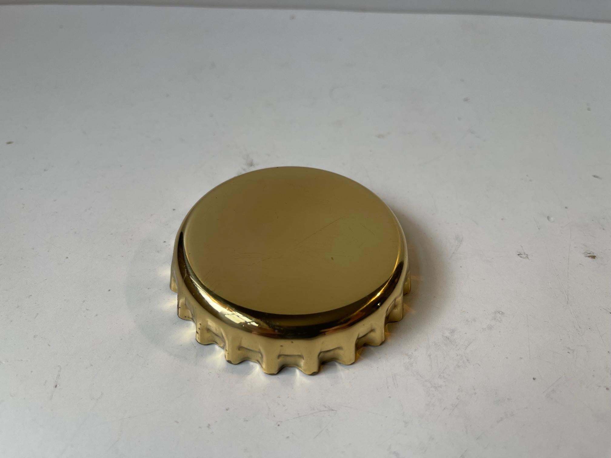 Manufactured in 1981 this curious bottle cap shaped opener is made from sculpted brass with a 'opener' mechanism in stainless steel. It is signed to the inside: Georg Jensen Design, Denmark, 1981.