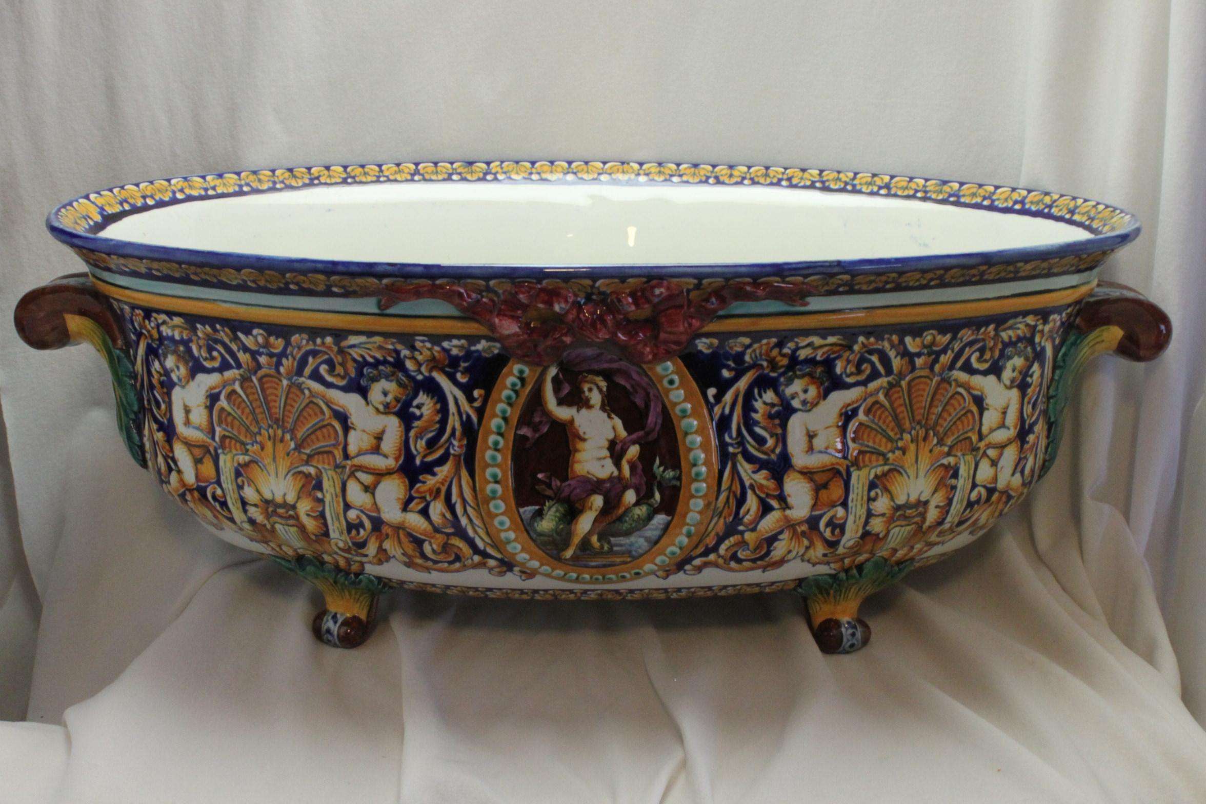This large, very colourful, twin handled Gien Faience jardiniere is hand painted and features a god and goddess on either side seated below red coloured bows and numerous cherubs supporting shell shaped fountains. It stands on four feet and measures