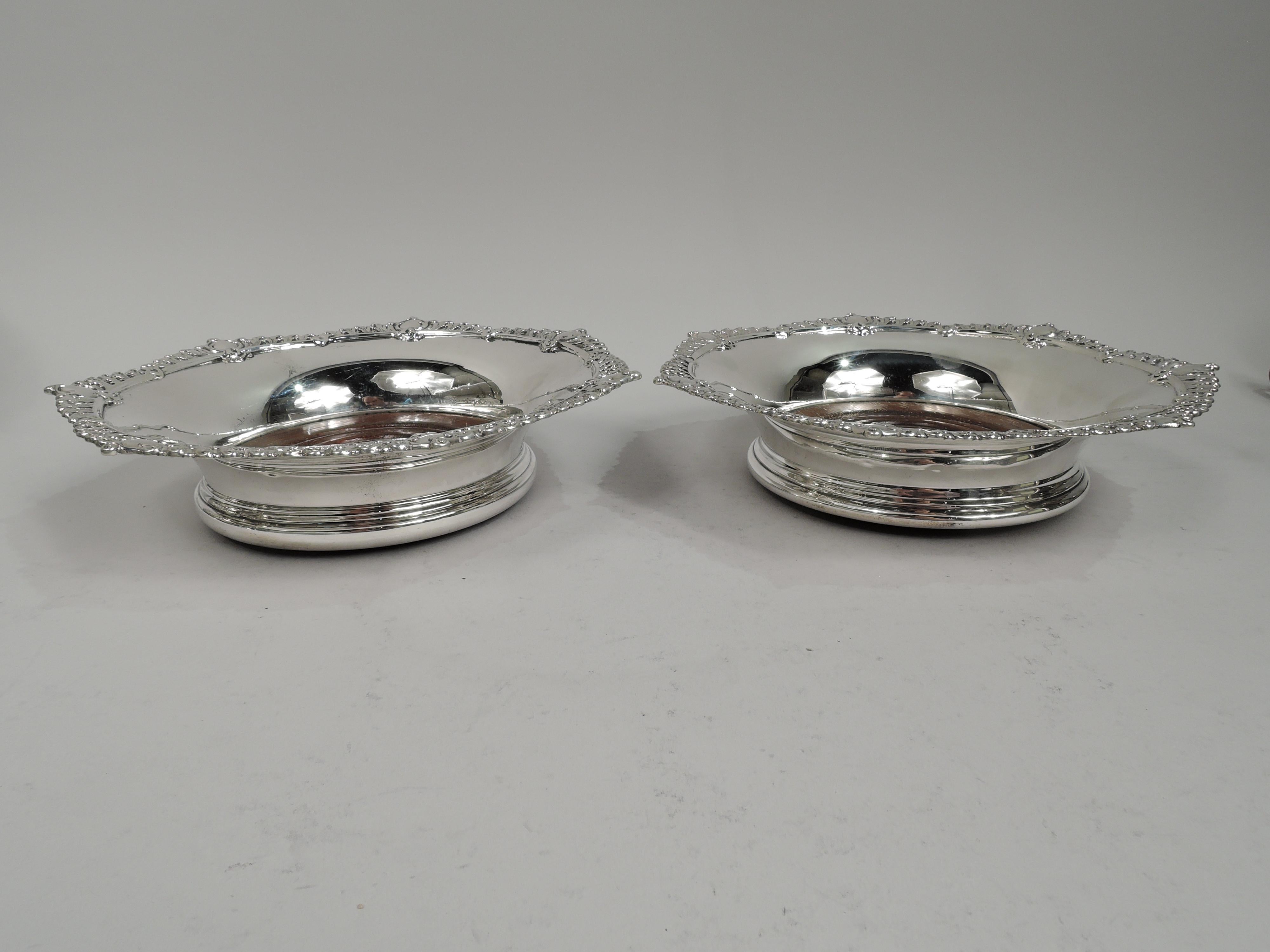 Pair of gift-quality American Edwardian Classical sterling silver wine bottle coasters, ca 1910. Retailed by Black, Starr & Frost in New York. Each: Tapering sides and spread and stepped base. Scrolling leaf rim interspersed with scrolled rondels.