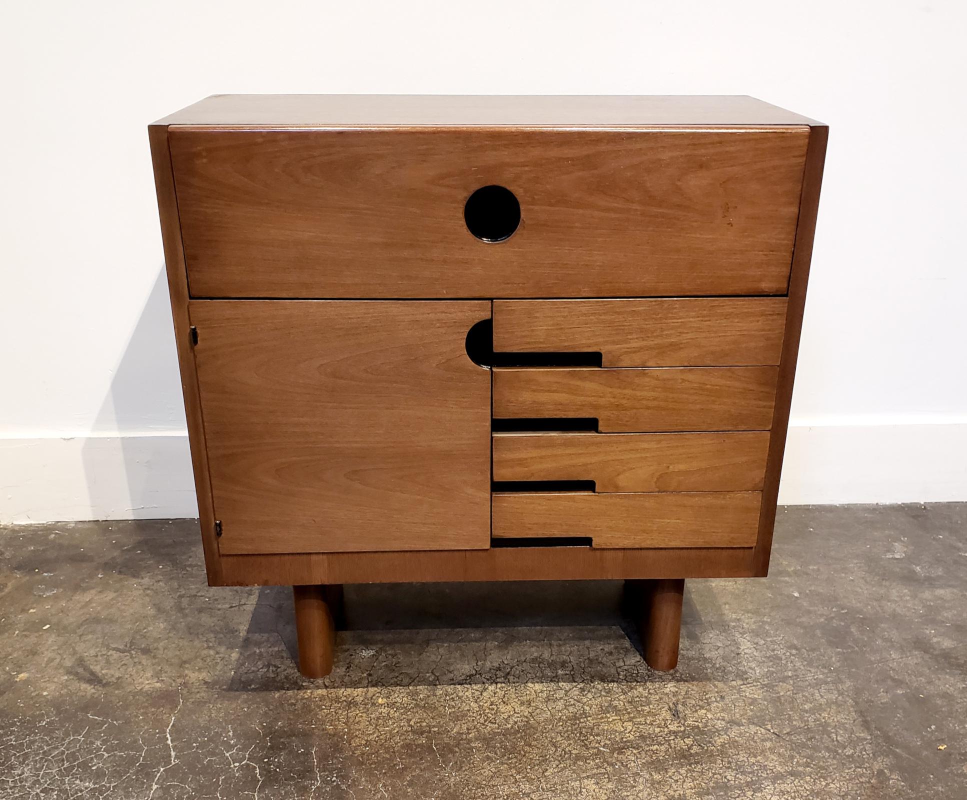 Modernist Art Deco era cabinet designed by Gilbert Rohde for Herman Miller circa 1930's-1940's. Sleek minimalist design in Mahogany wood with pulls accented in black. Top pulls down to reveal several small hutches and trays for papers/letters, etc.