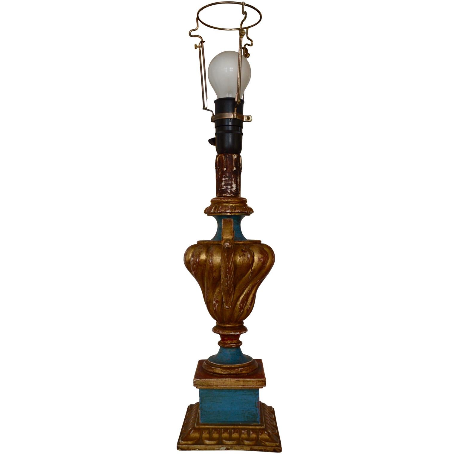 This 19th century Rococo-style, gilded and painted in blue and red table lamp, featuring an decorative urn shaped body with side handles carved in wood. The lamp is raised on a square base.