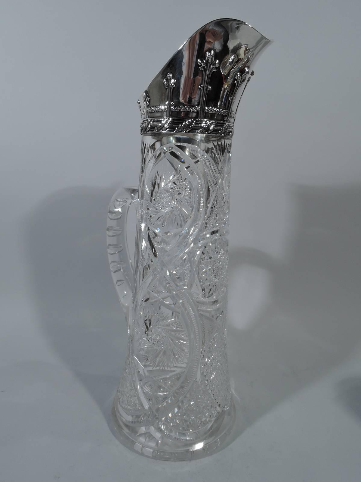 Gilded Age brilliant cut-glass claret jug with sterling silver collar. Made by Tiffany & Co. in New York. Tall and upward tapering with c-scroll handle. Stars, diaper, flowers, and ferns set in interlaced scrolled frame. Collar has helmet mouth with