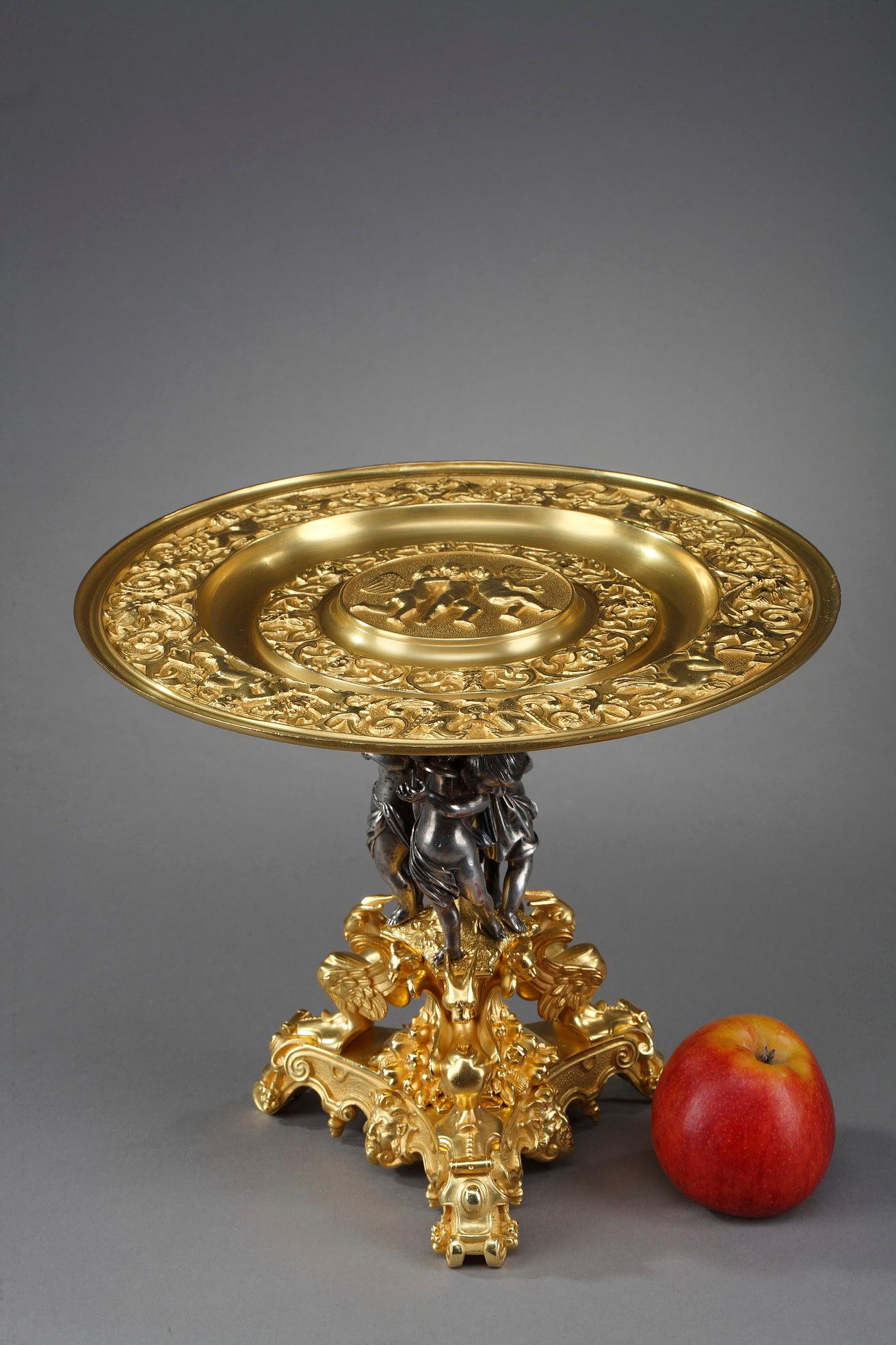 Centerpiece in gilt bronze decorated with three putti musicians in silver bronze on the shaft. The central tray in gilt bronze is engraved with motifs and three putti in the center. It rests on a base decorated with three winged griffins and heads