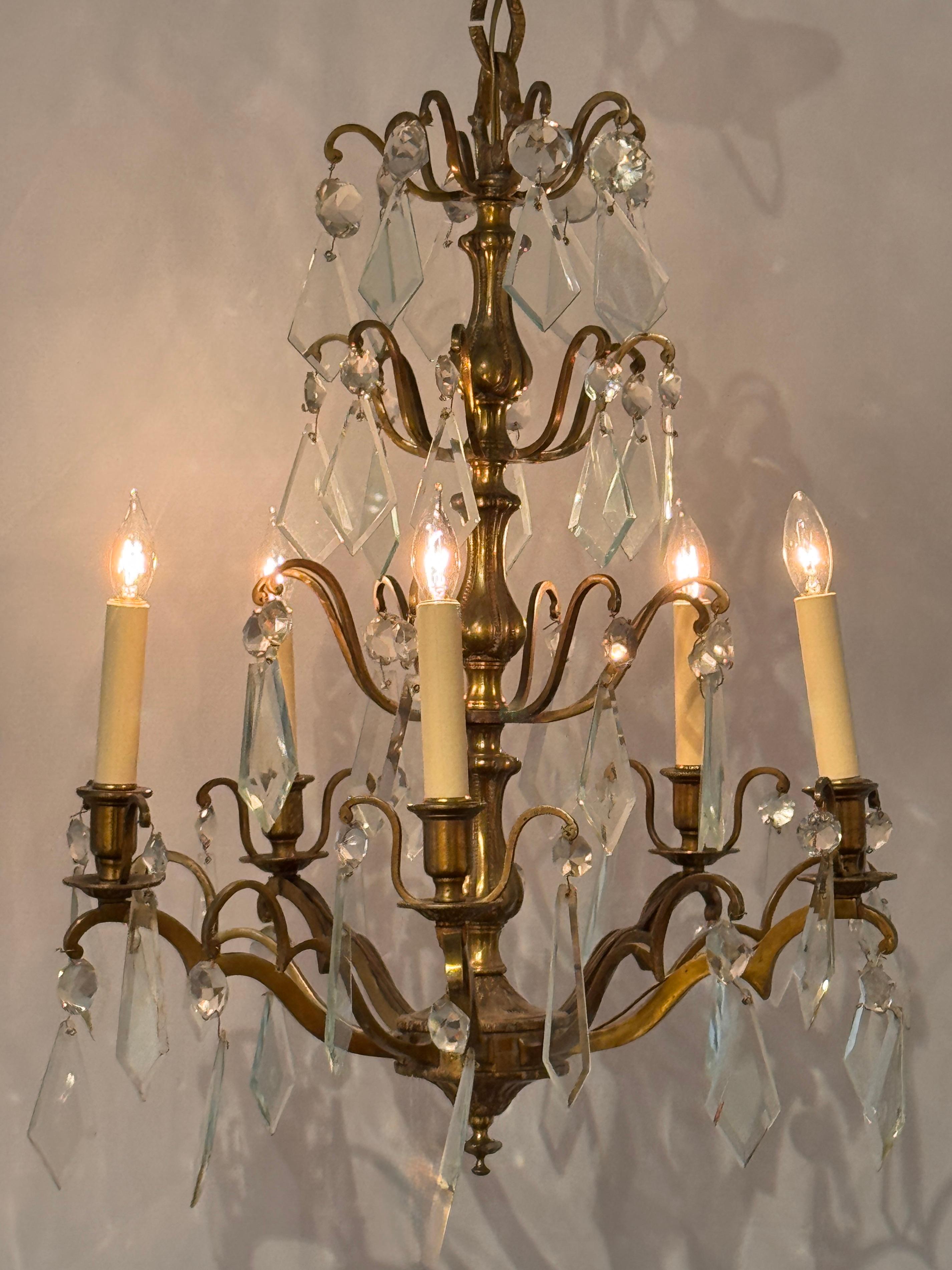 Six-arm chandelier in gilded bronze with elegantly scrolled arms and a tiered center column. Features faceted round and diamond-shaped crystal drops throughout, suspended beneath each arm and along the upper tier. The warm gilded surface contrasts