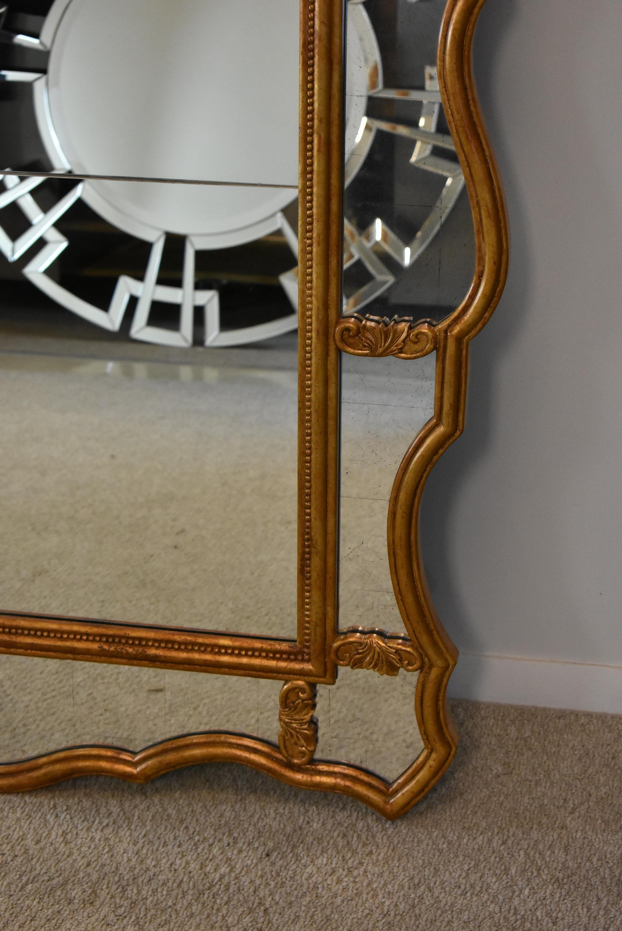 Gilded floor mirror by John Richard. Mirror features a serpentine arched top with a paneled boarder throughout. Each section has a distressed mirror with a thin beveled edge. Carved wood frame has a gilded bronze gold finish.