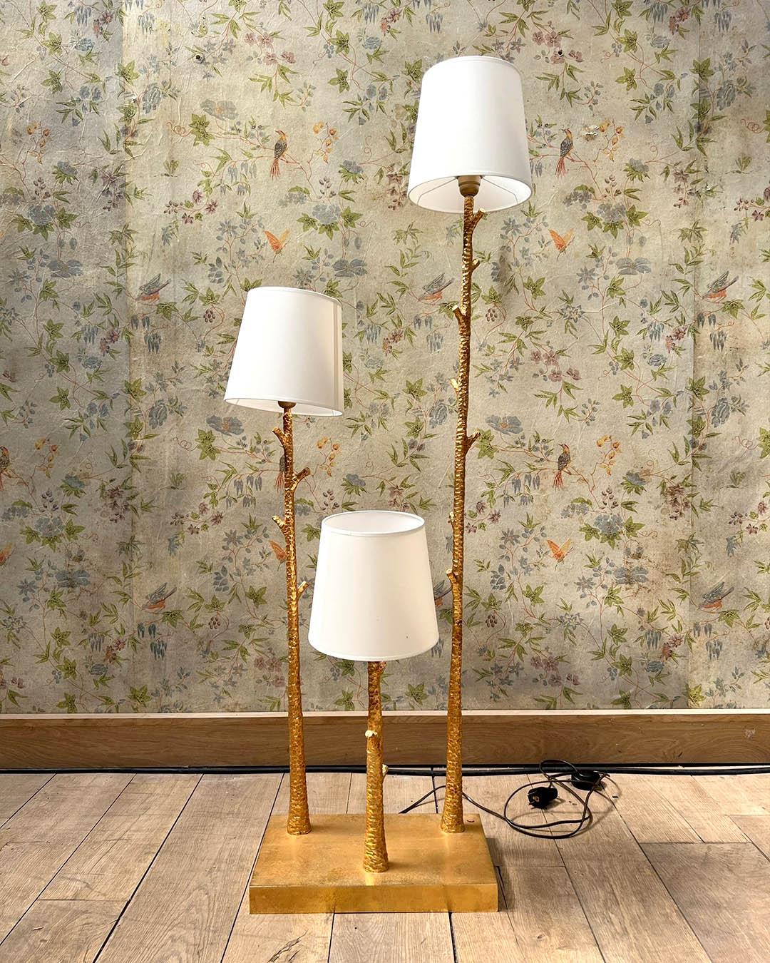 A striking floor lamp crafted in hammered gilded metal, designed with three sculptural branches rising at varying heights, each topped with a white conical lampshade. The textured gold finish gives the illusion of tree bark, adding a naturalistic
