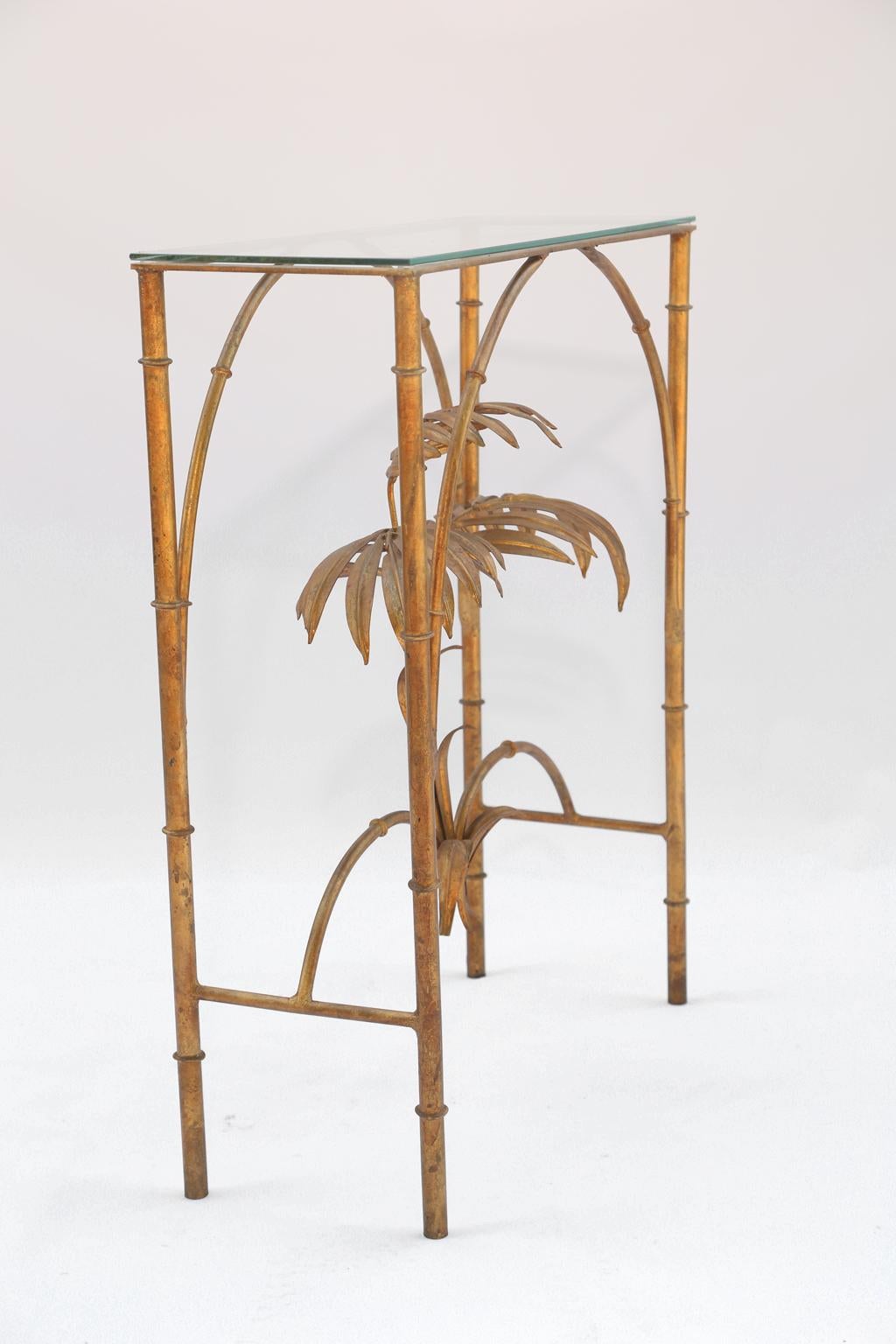 Console, of iron, in the Hollywood Regency style, having a gilded finish, its rectangular glass top on faux bamboo table base of four legs, joined by double arches, framing a palm tree.

Stock ID:  D1824