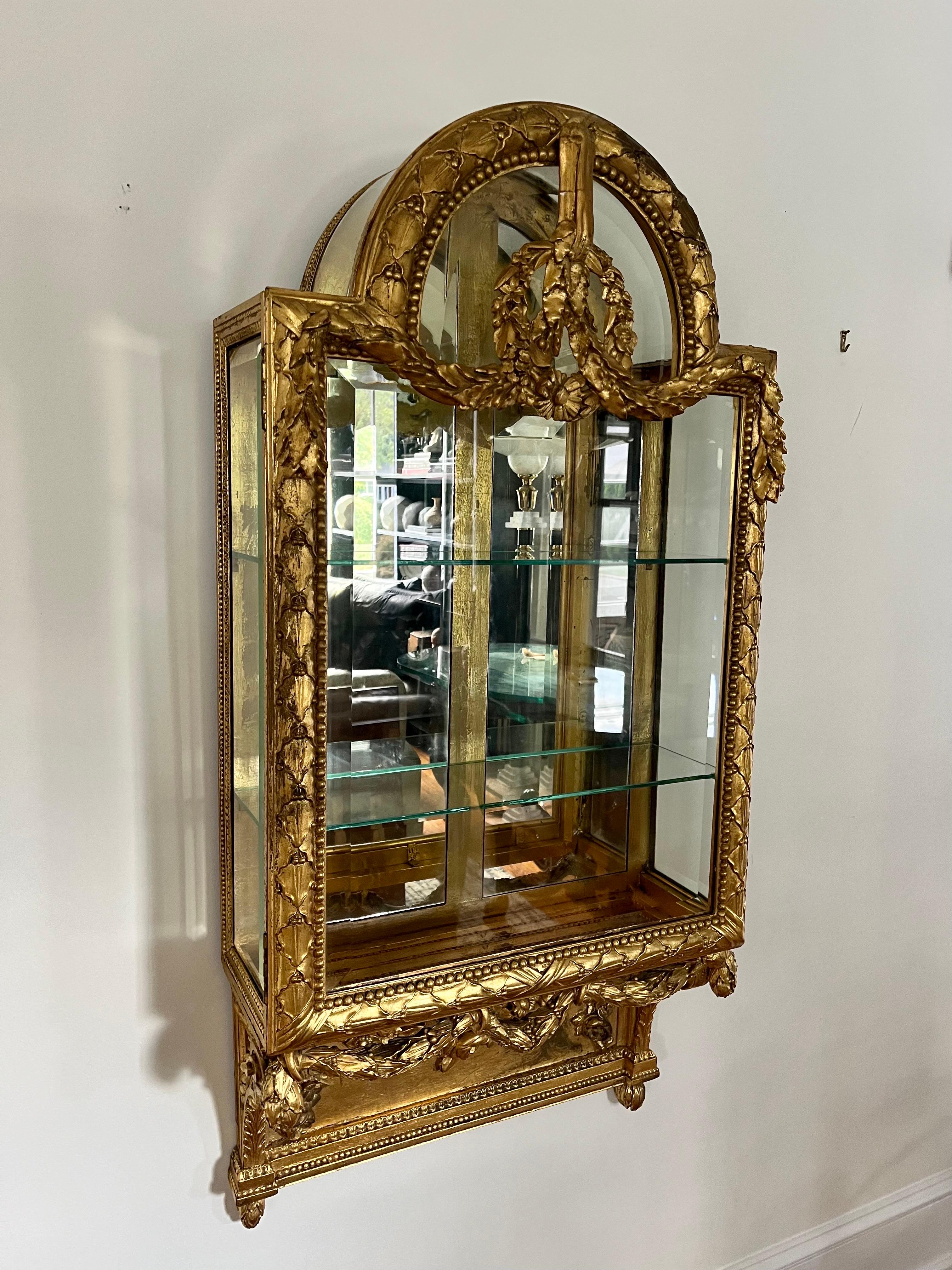 Beautiful wall hanging Vitrine in the style of Louis XVI.  Elaborate decorative french Louis XVI style giltwood front with beveled glass and antiqued mirror interior and 2 glass shelves. Sides and top are decorative metal and cabinet has 2 skeleton