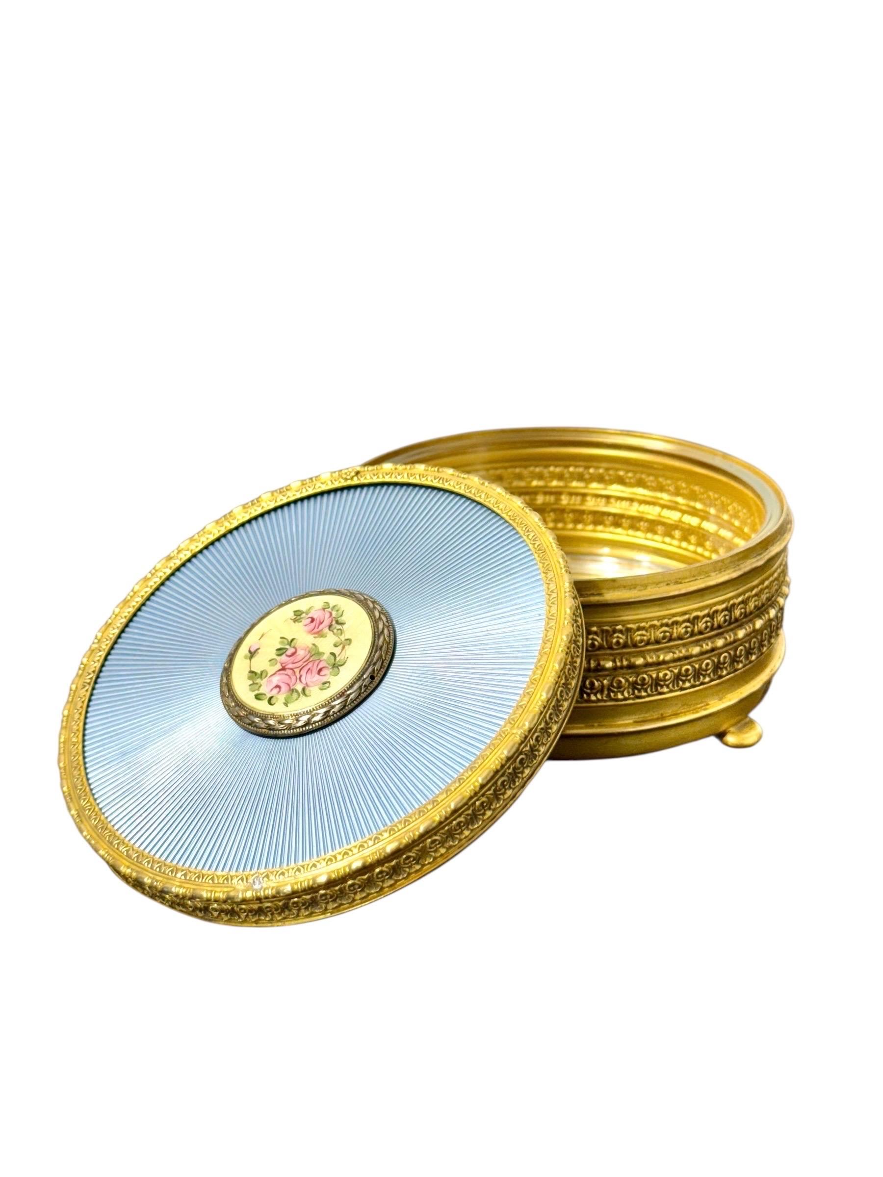 Cette boîte à bijoux exquise combine un travail de métal raffiné, un émail guilloché délicat et un élégant contenant intérieur en verre, offrant à la fois beauté et praticité. Le couvercle est sa caractéristique la plus frappante : une surface
