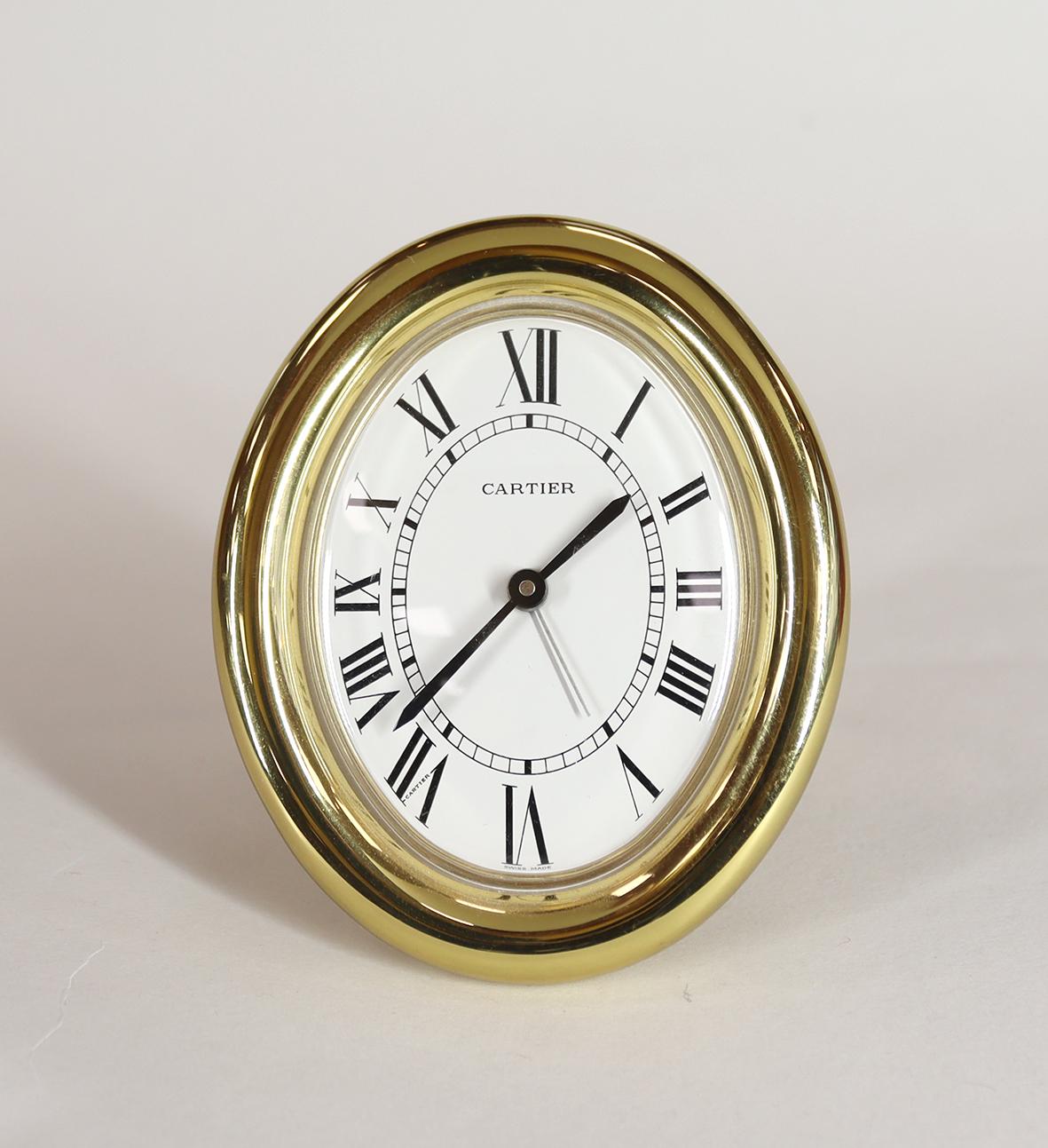 A Cartier Les Must Alarm Clock in an oval gold plated frame, with cabochon jewelled setting nuts, resting on a strut back with the distinctive Cartier ‘C’ forming the feet.

This Swiss made quartz clock stamped ‘Cartier a Paris’ comes with it’s