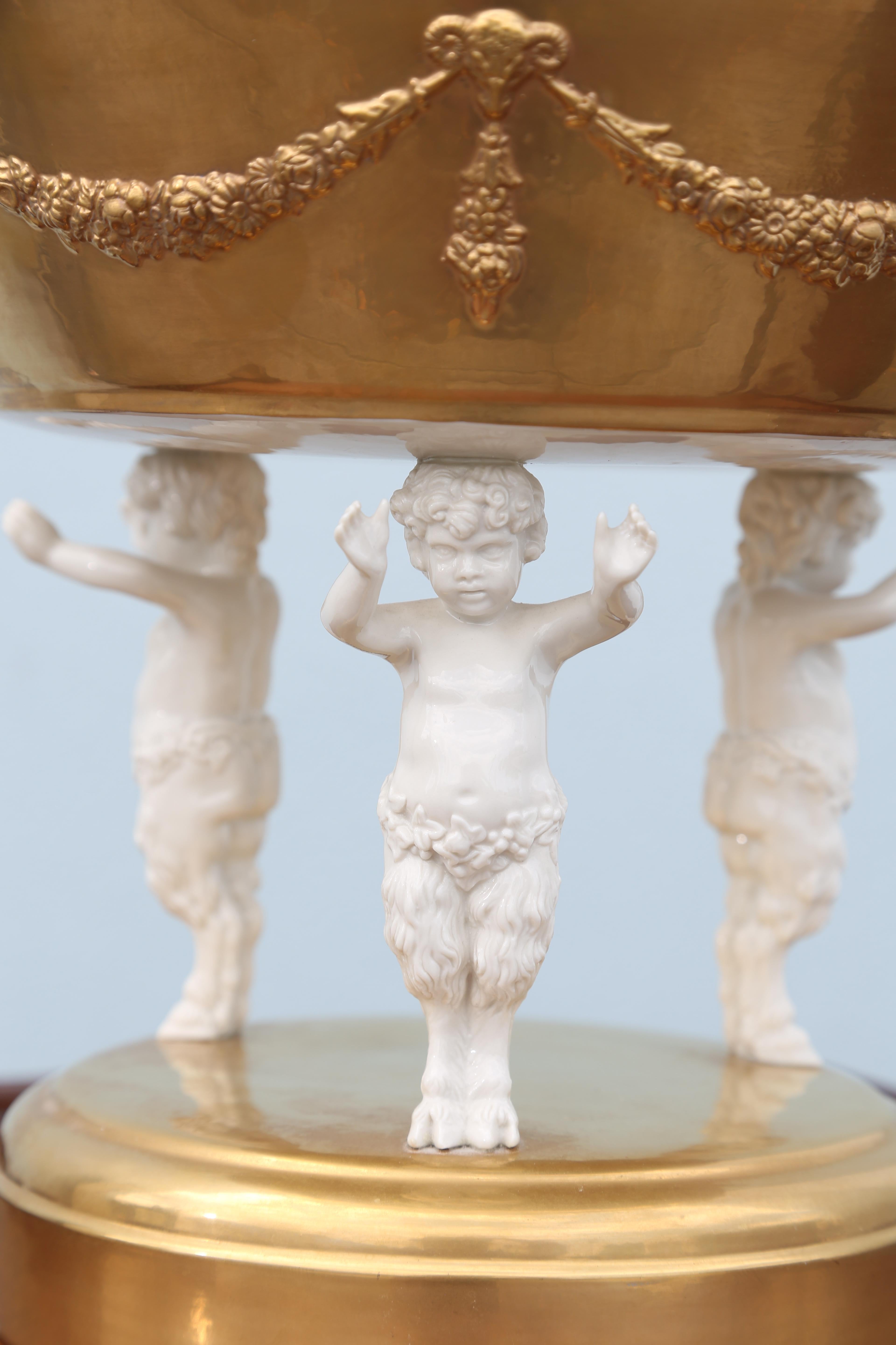 Centro de mesa de porcelana dorada con tres putti mitológicos de pie sobre una base redonda que sostienen un gran cuenco sobre sus cabezas.