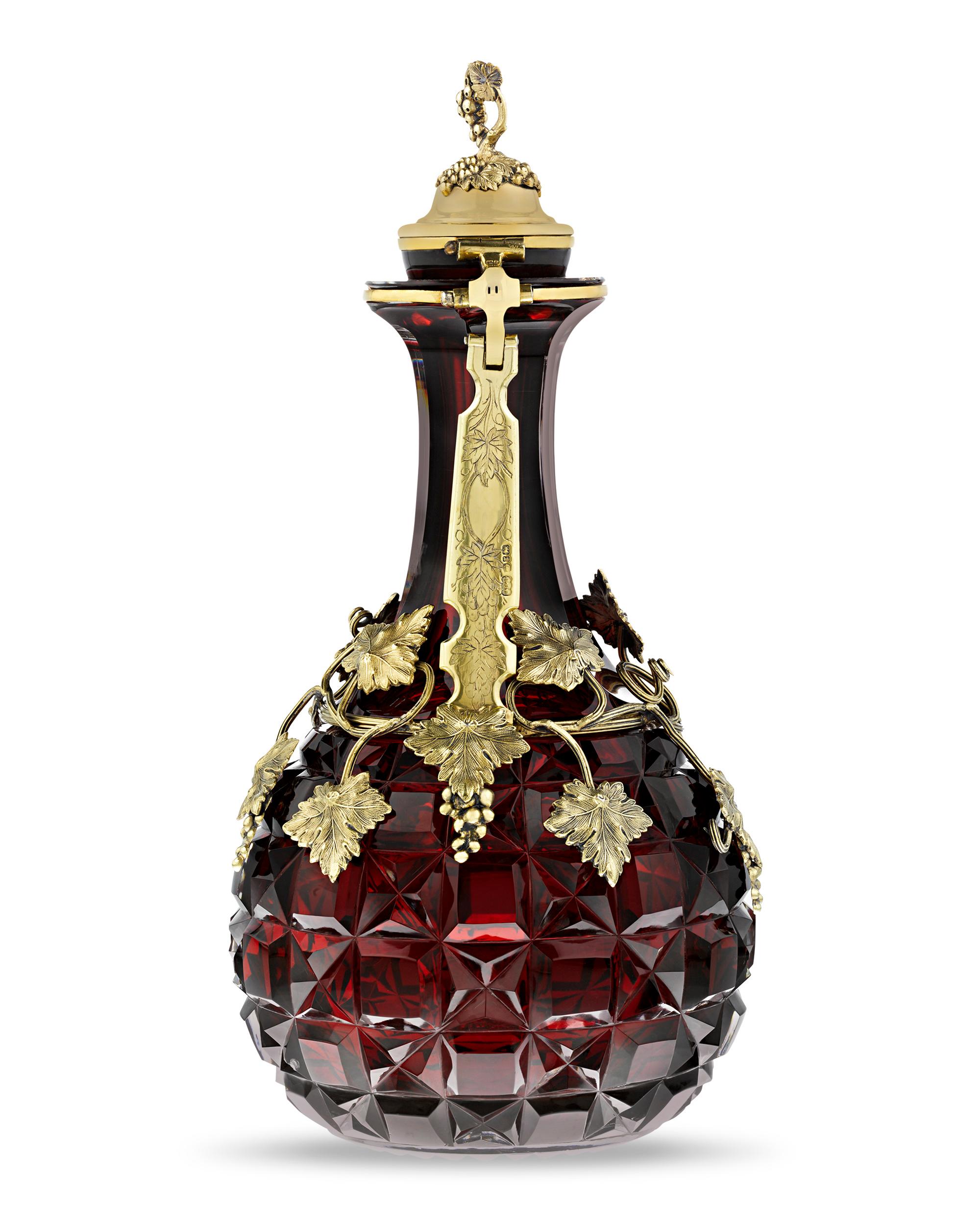This pair of stunning cut cranberry glass jugs are mounted with gilt silver in a grapevine motif, indicating their intended use as wine decanters. The vibrant red glass vessels feature deep, precise cutting of fine caliber while the silverwork is