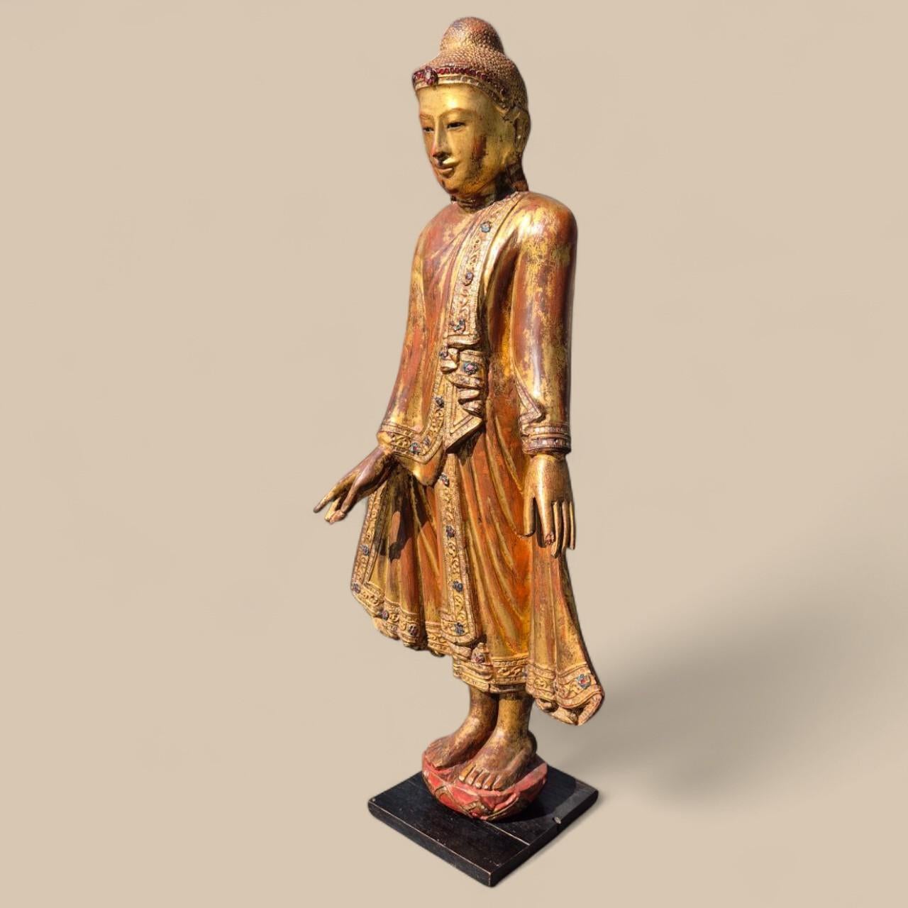 Superb statue of a young standing Buddha of Burmese origin, hand-carved in teak wood, gilded with gold leaf, and adorned with fine inlays of colored stones.

His delicately sculpted face and serene gaze give him a powerful spiritual and calming