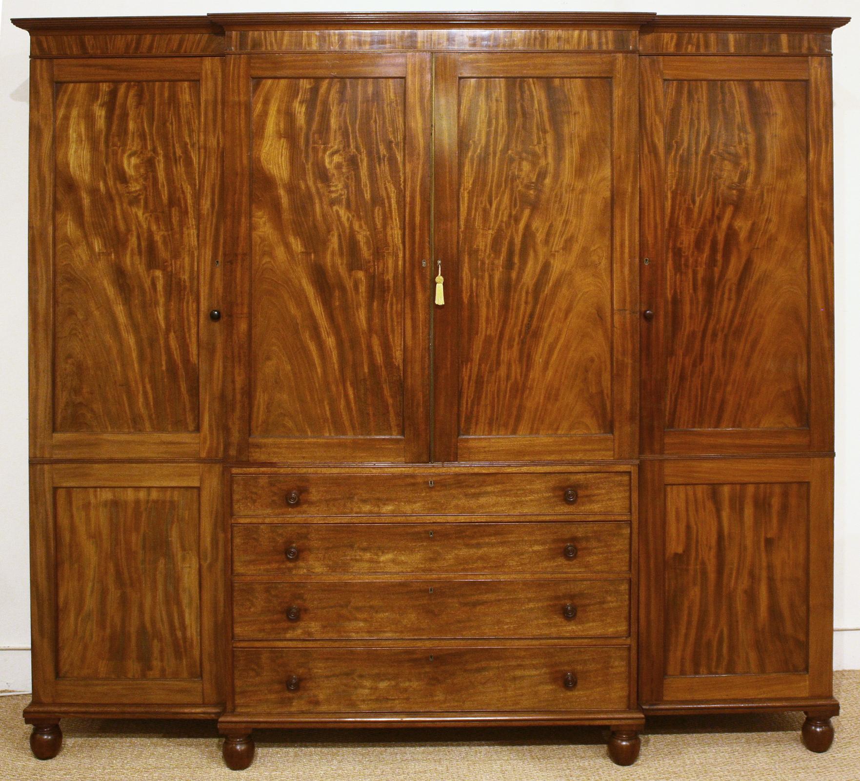 A stunning quality Georgian Revival breakfront mahogany gentleman’s wardrobe/press. The cornice has a broken pediment with a carved laurel wreath in center and two inlaid garland motifs. The central twin doors open to reveal five oak dressing trays