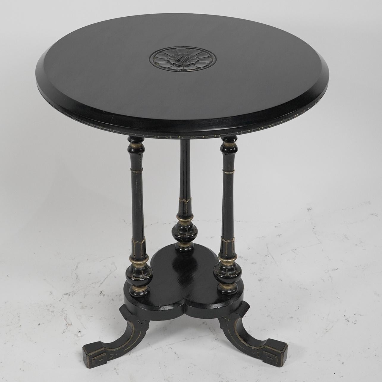 Gillows attributed. An Aesthetic Movement ebonized circular side table with carved sunflower floret to the top with three turned, carved and incised gilt decoration to the legs and to the splayed feet which unite to a trefoil floret lower shelf.