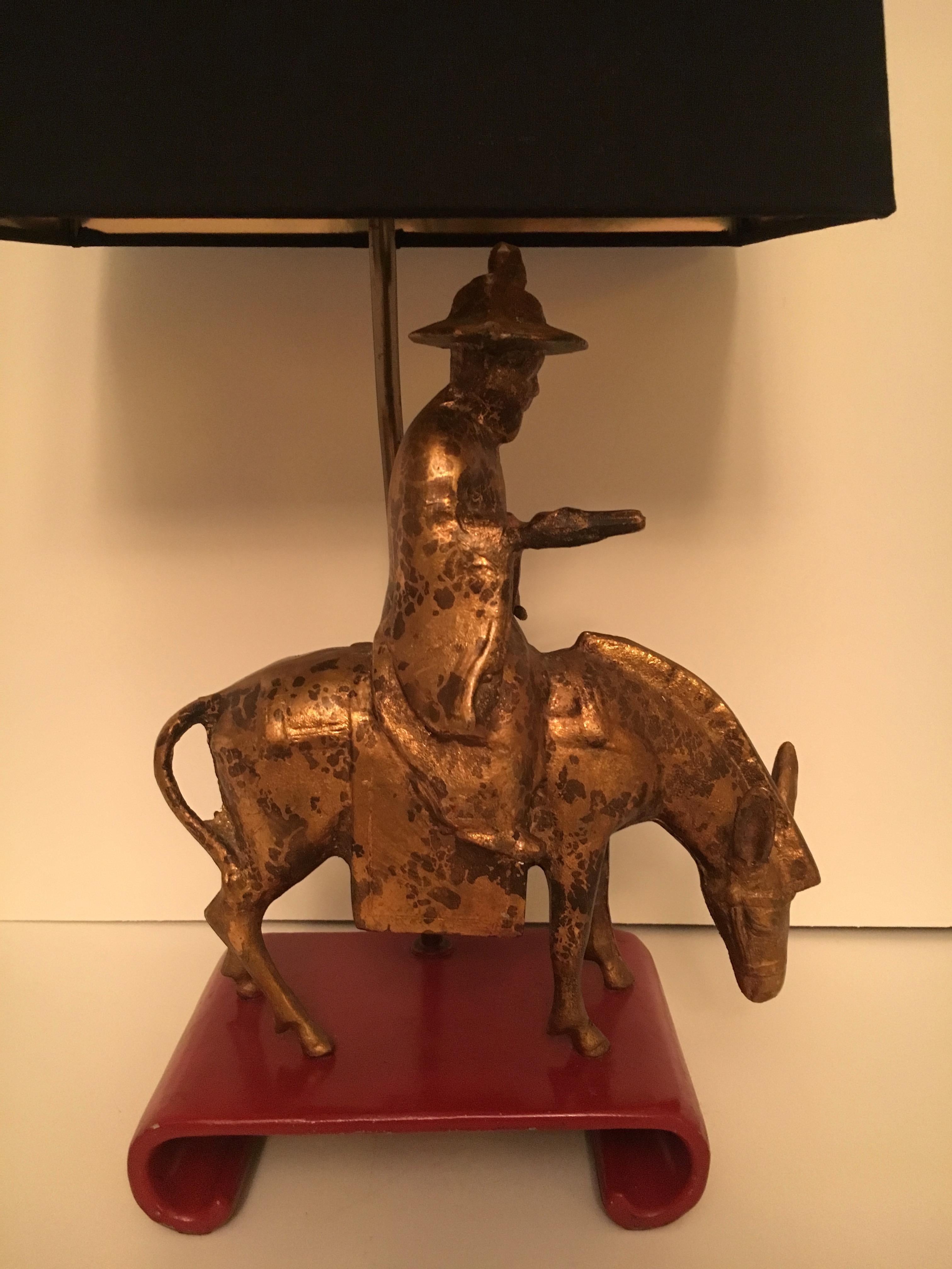 Gilt Asian figure on a horse or mule displayed a red lacquered Asian platform. The lamp is extremely handsome and with a very good energy. We have placed a 'black out' shade lined in gold for atmosphere lighting. The finial is a carved resin or bone