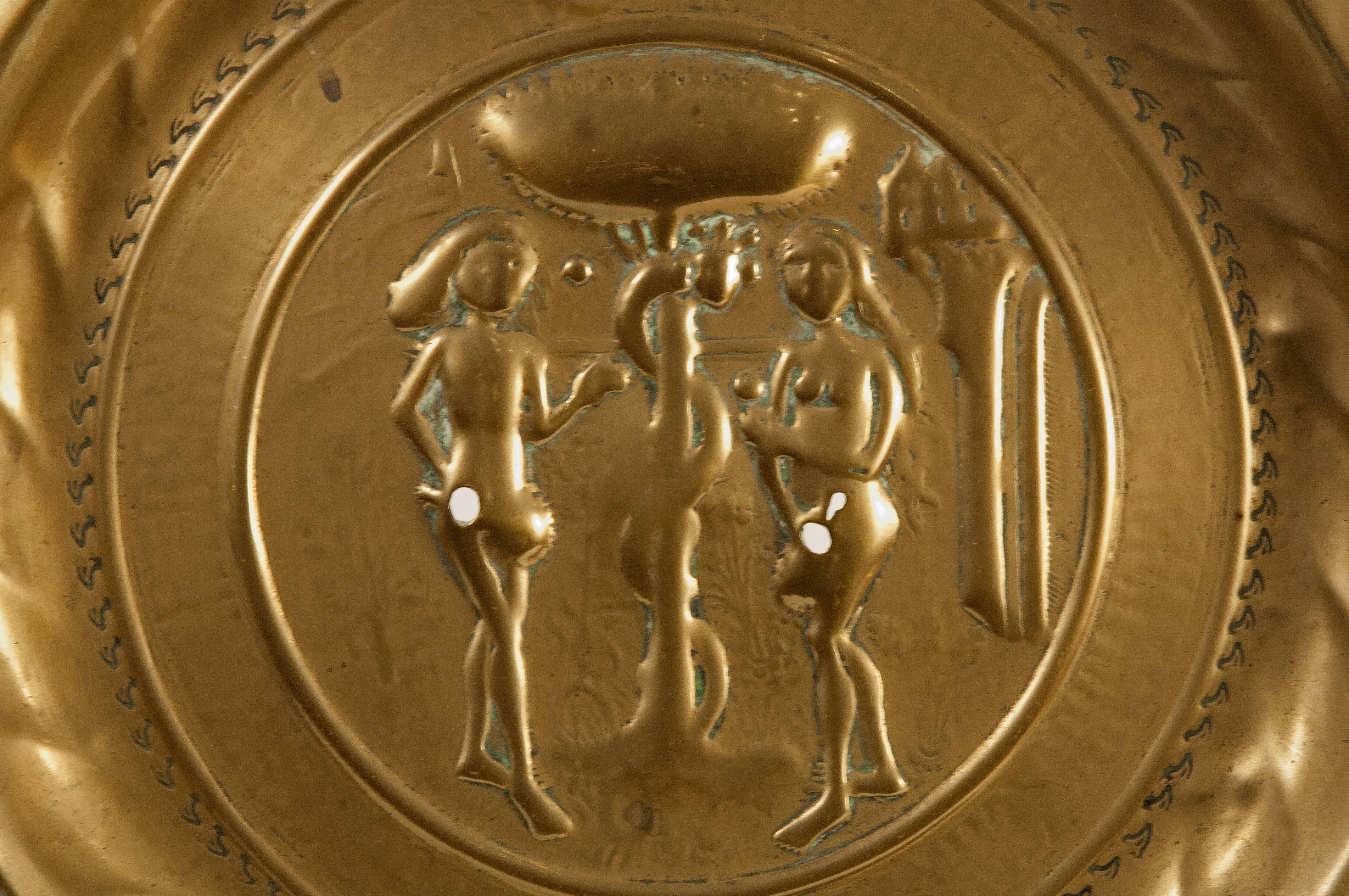 Alms collection dish made of gilded brass decorated with a series of moldings and details both on the edge and towards the center. In this area there is a figurative motif in light relief (Adam and Eve flanking the tree in which the Serpent of