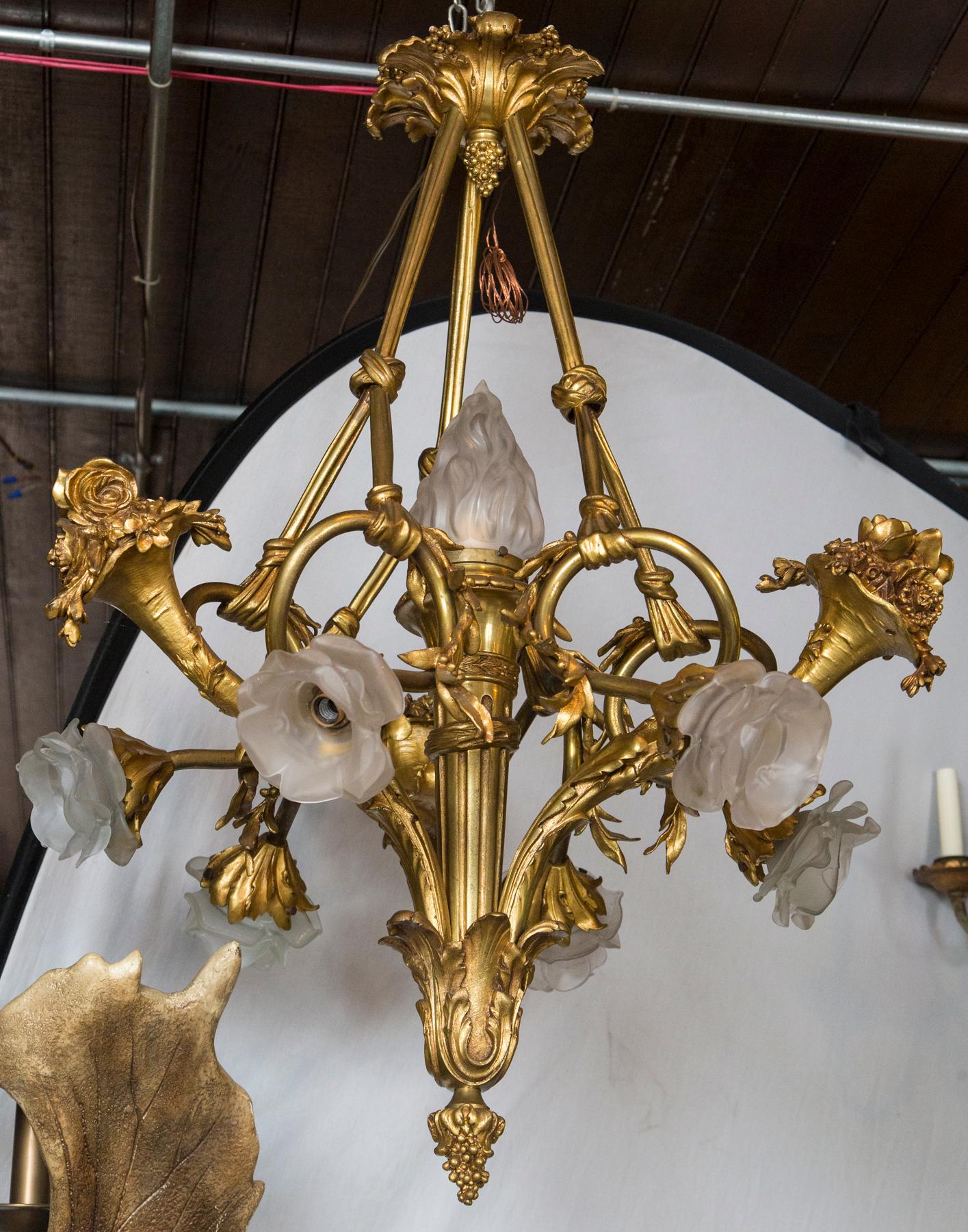 The central torch form topped by a frosted glass flame shade. Six other frosted glass floral form shades turn downward. Acanthus leaf for candle cups, with acanthus form decoration on the lower part, the arms, and the ceiling cap. A grape form
