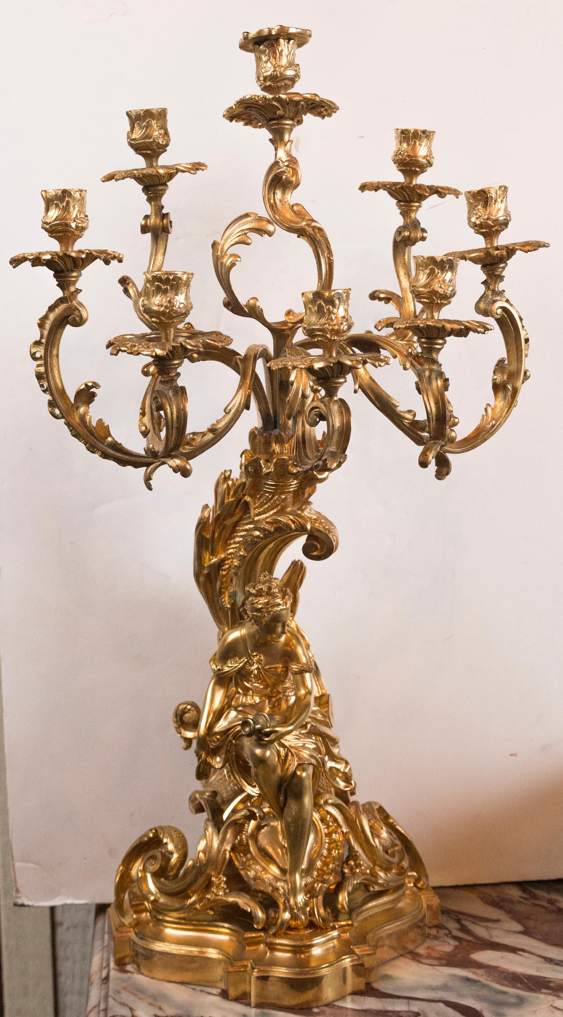 Made in 2 parts. The candelabra fits into the large candle cup above the seated female figure. She holds a wine jug at her knee in her left hand. She wears a loose fitting covering and sits upon scrolls of the Louis XV/Rococo style. The entire