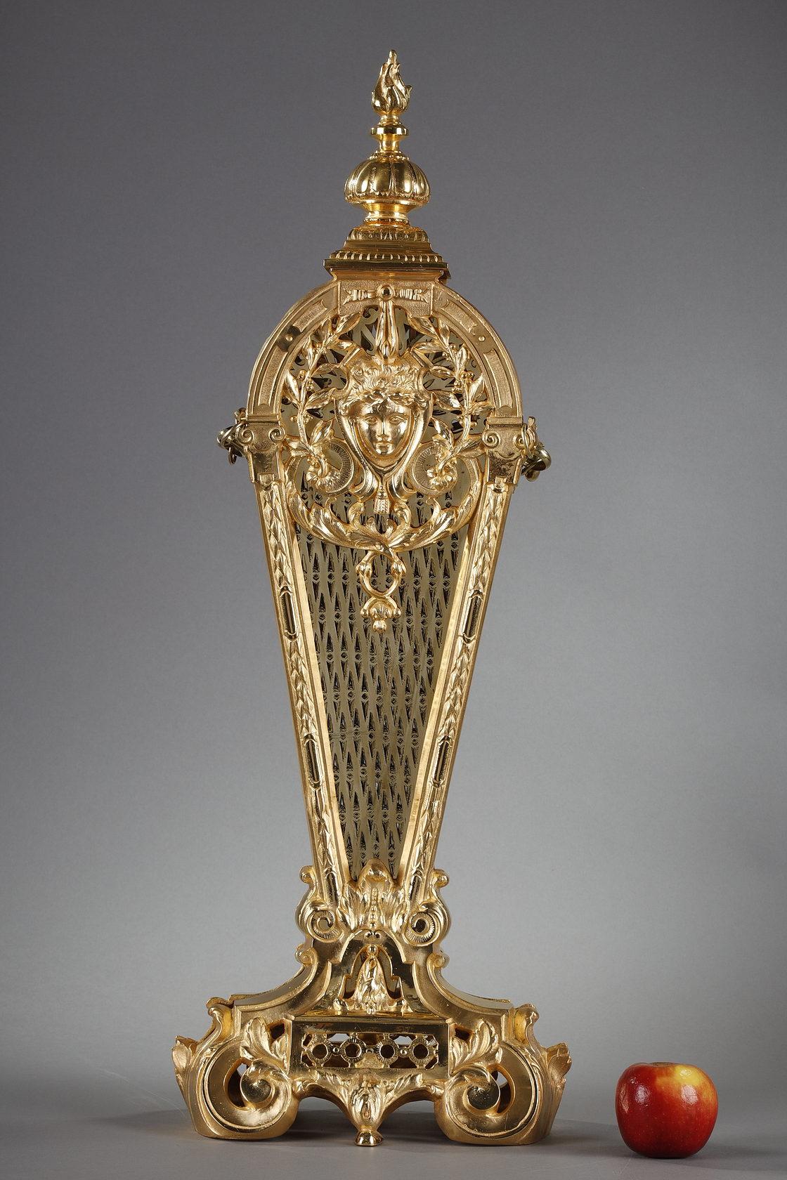 Fan-shaped mantel screen in brass and chased and gilded bronze. The central post is decorated with a female face surrounded by olive branches, trimmings and interlacing acanthus leaves. The fireguard is surmounted by a flame, a reference to the