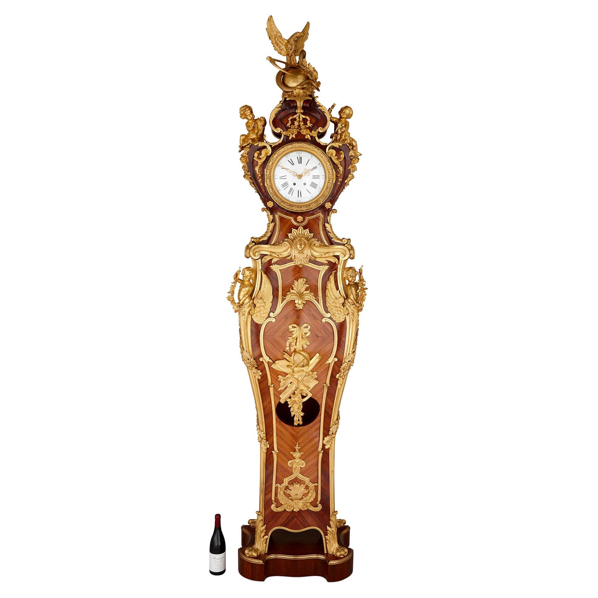 Gilt bronze and parquetry standing clock by Kahn in the Régence style
French, early 20th century
Measures: Height 262cm, width 62cm, depth 17cm

This beautiful longcase clock is of elongated violin body form. The wooden surfaces throughout are