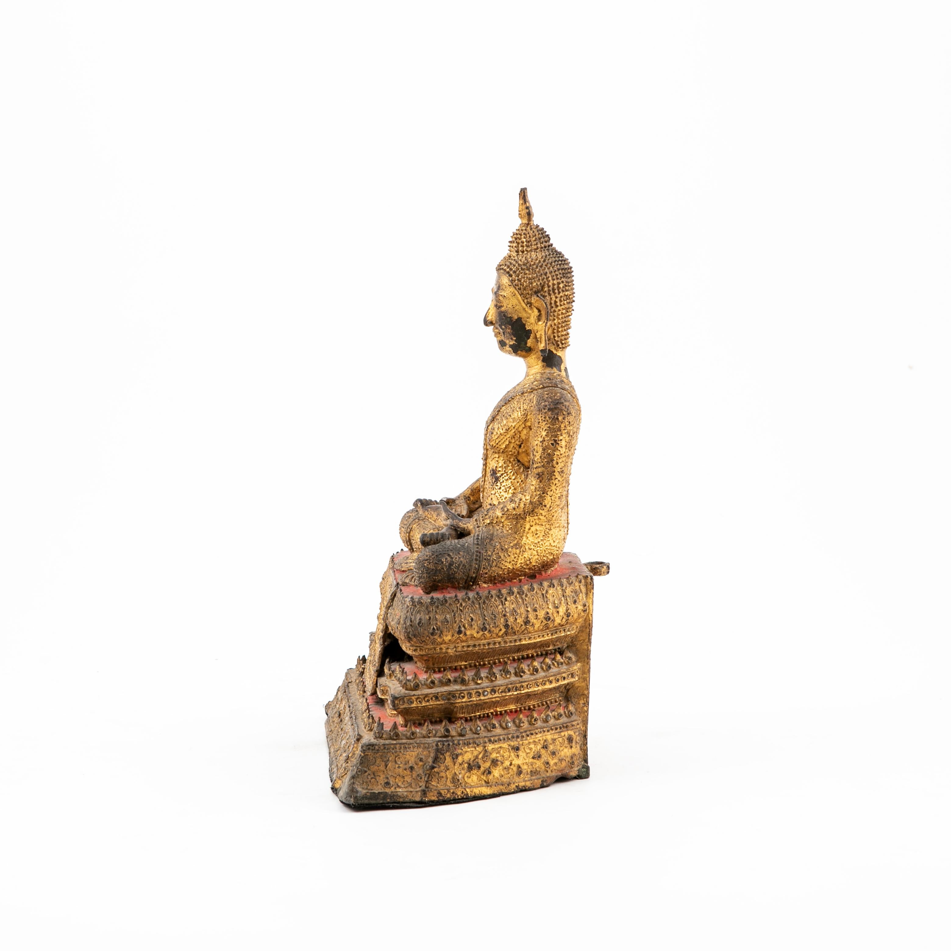 Bouddha assis du Siam en bronze doré avec des détails en laque rouge.
Les yeux fermés et les mains reposées indiquent la méditation.
Le Bouddha est assis sereinement en position de demi-lotus.
 
État original intact avec une belle patine naturelle