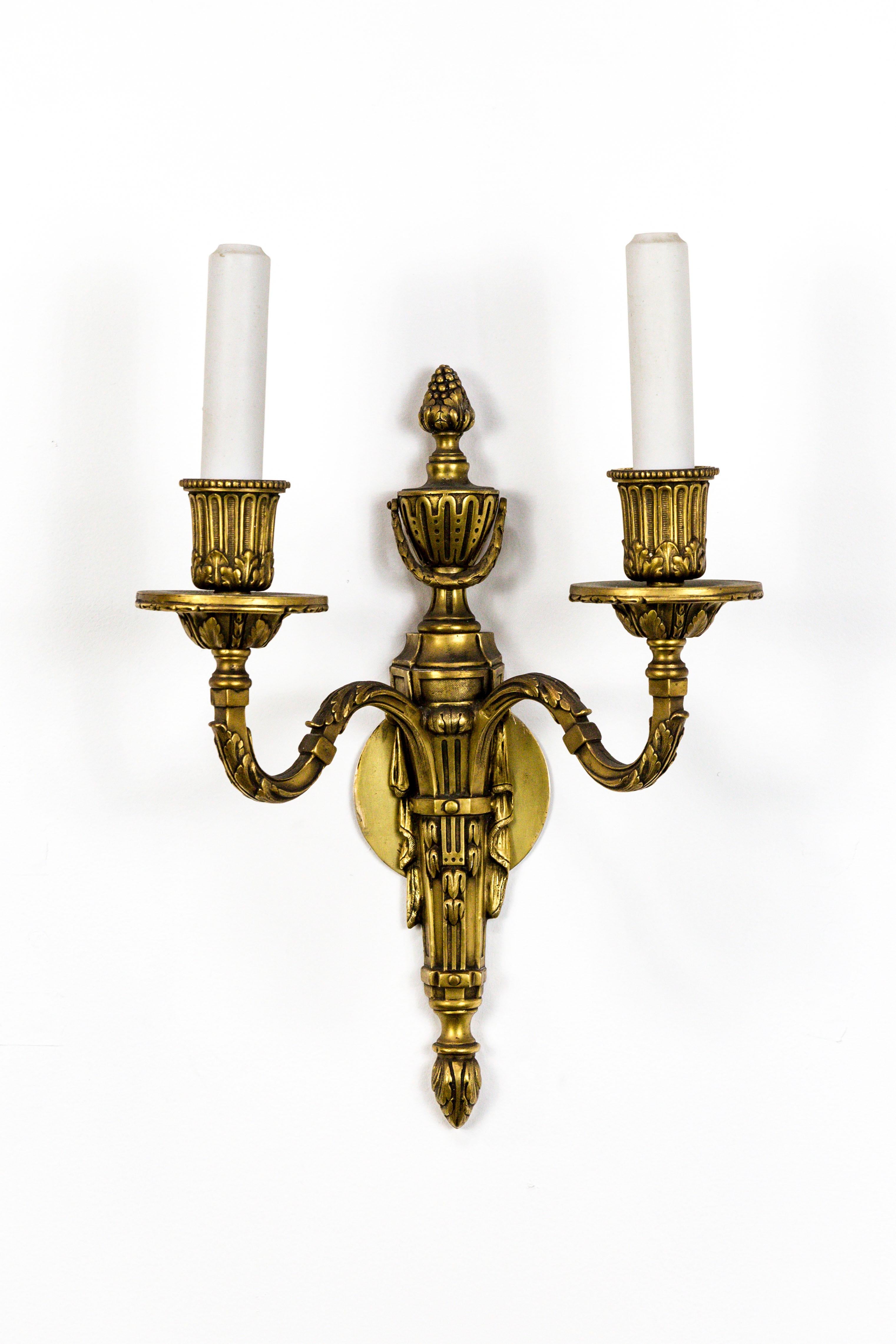 Regal, 1940s, two-light Empire style candelabra sconces in gilt bronze with ceramic candle covers. Adorned with acanthus leaf swag, flora and reed detail, and urn and acorn top. American. Measures: 15.5” height x 10.5” width x 5.75