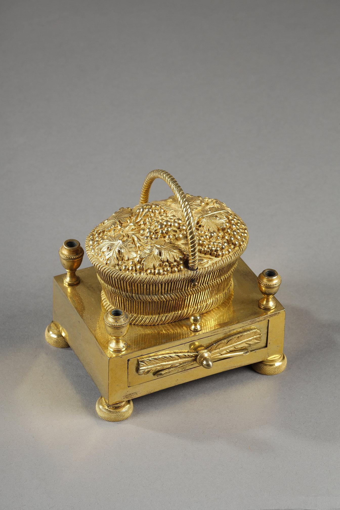 Gilt bronze inkwell, concealed in a grape basket, revealing two cups. The rectangular base is adorned with four urns at the corners, acting as nib holders, and with an applied semi-circular rotating drawer.

